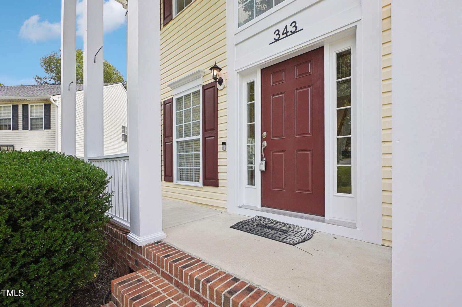 The image shows the entrance of a house with a maroon front door and white trim, accented by a black welcome mat. The house features yellow siding and white columns, with a small brick step leading to the porch. A hedge borders the walkway.