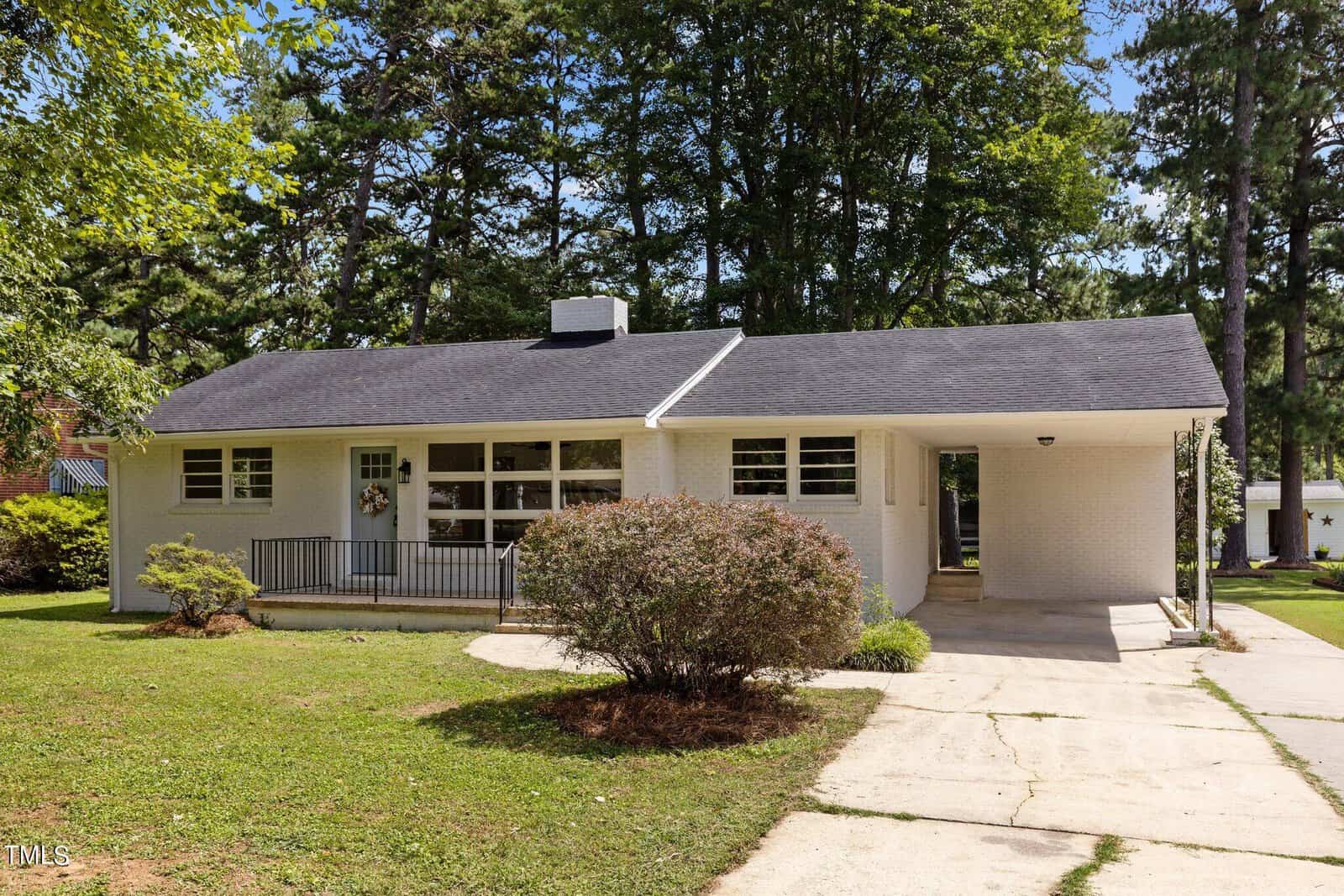 Nestled at 215 Pine Tree Road, this charming Oxford home is a white single-story retreat with a dark roof. Encased by trees, it boasts large windows, a welcoming front porch with railing and an attached carport. A driveway leads to the house, framed by shrubs and a lush green lawn.