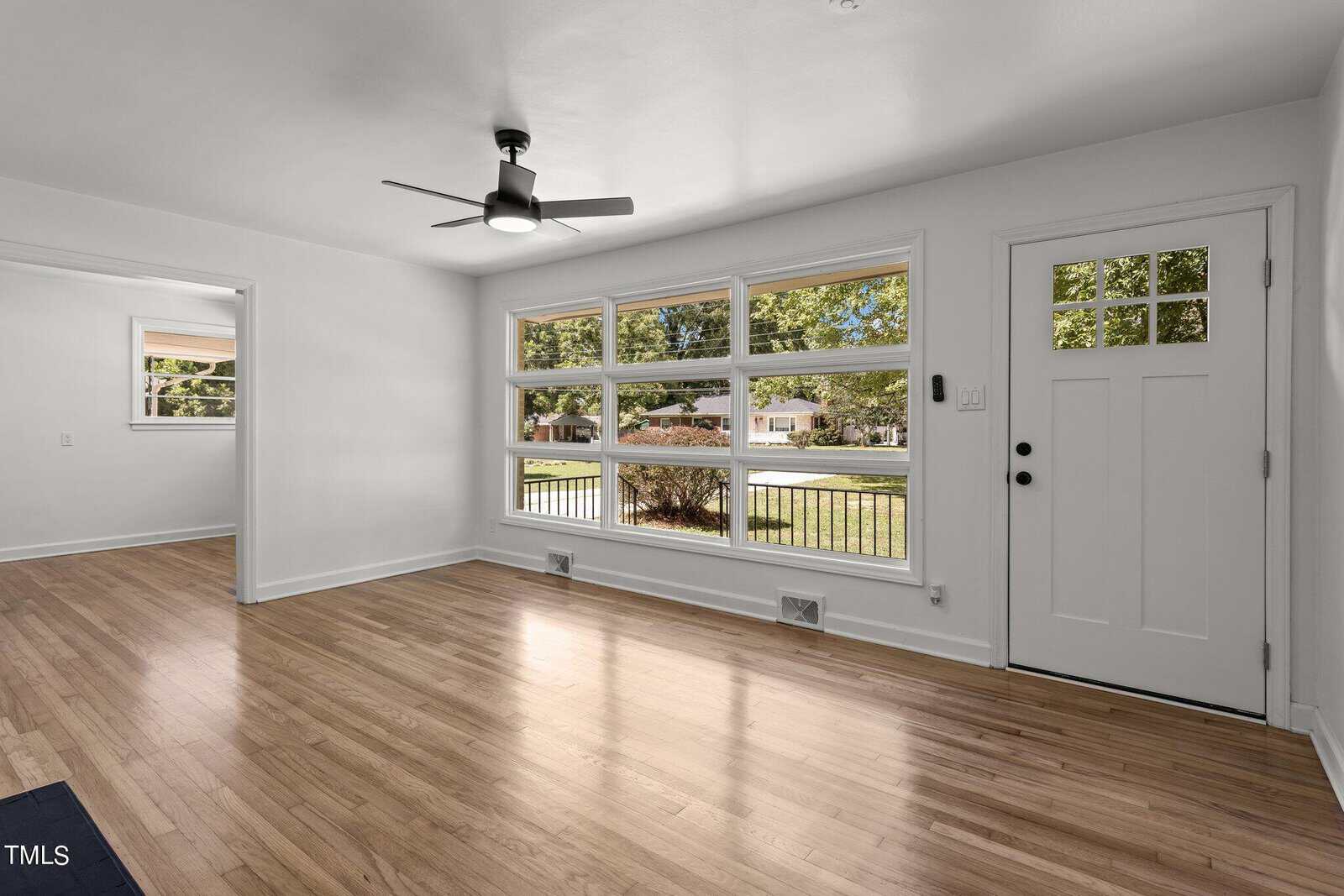 Bright living room with large windows, light wood flooring, and a ceiling fan. The space opens to a room on the left, while a white front door with glass panels is on the right. Outside, greenery is visible through the windows.