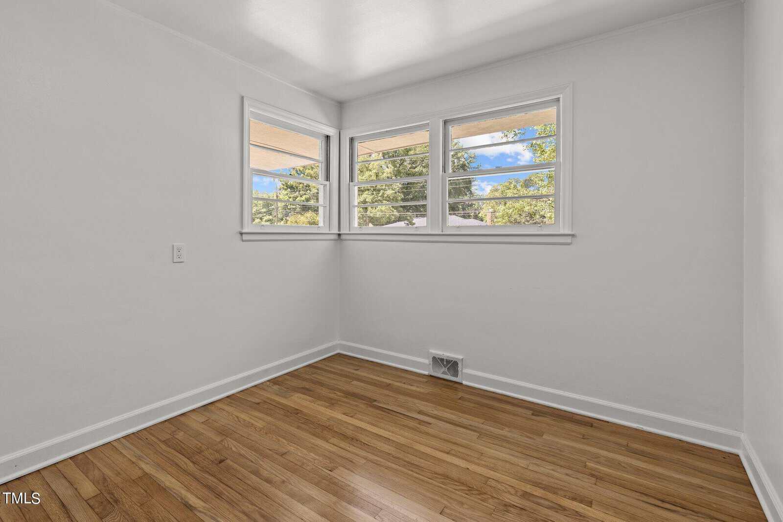 A small, empty room with light wooden flooring and white walls. Three rectangular windows on one wall let in natural light, offering a view of trees outside. The room has a clean, minimalist appearance.