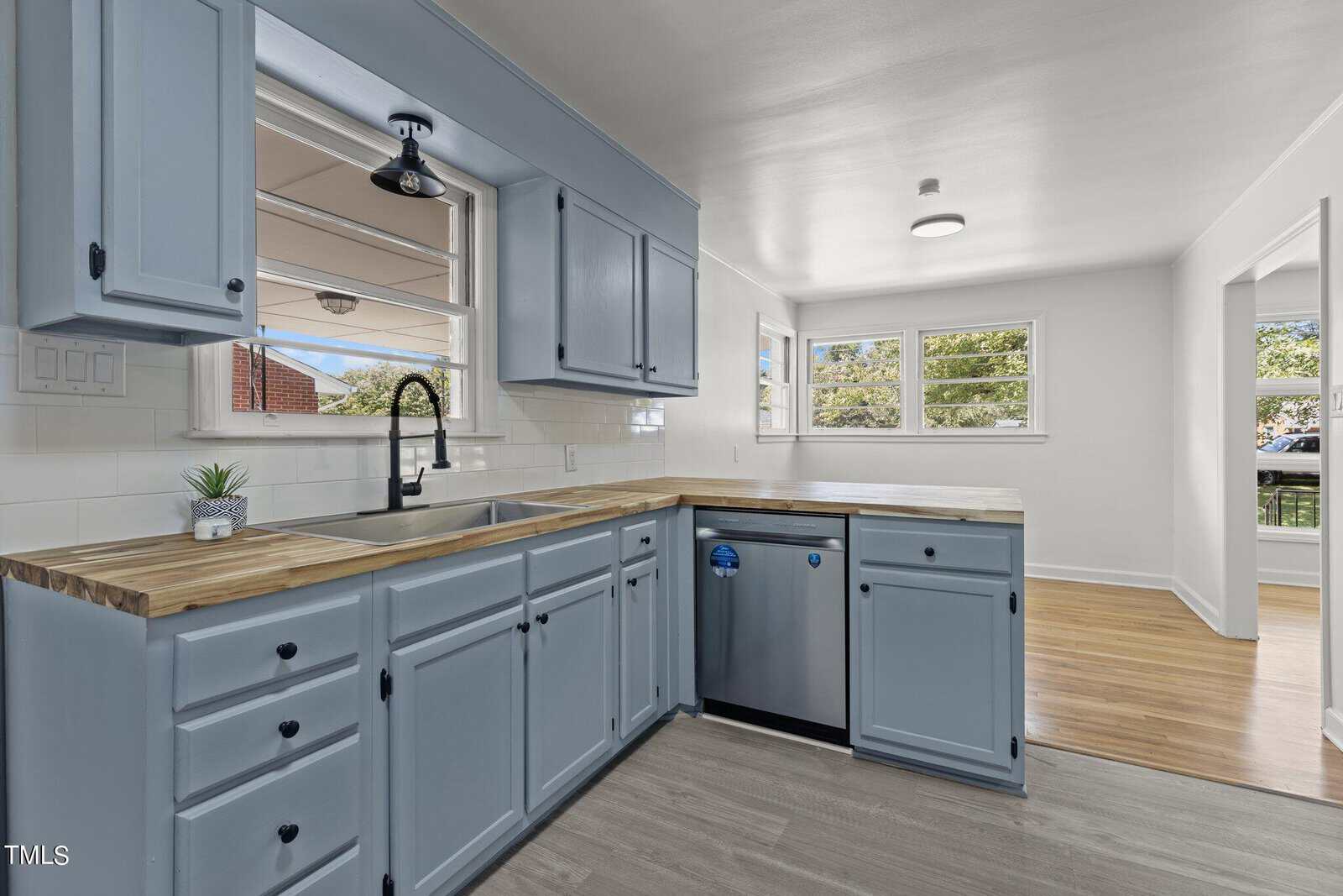 A modern kitchen with light blue cabinets, wood countertops, and stainless steel appliances. The room features a large window with natural light, a potted plant by the sink, and a wooden floor extending into an adjoining room.