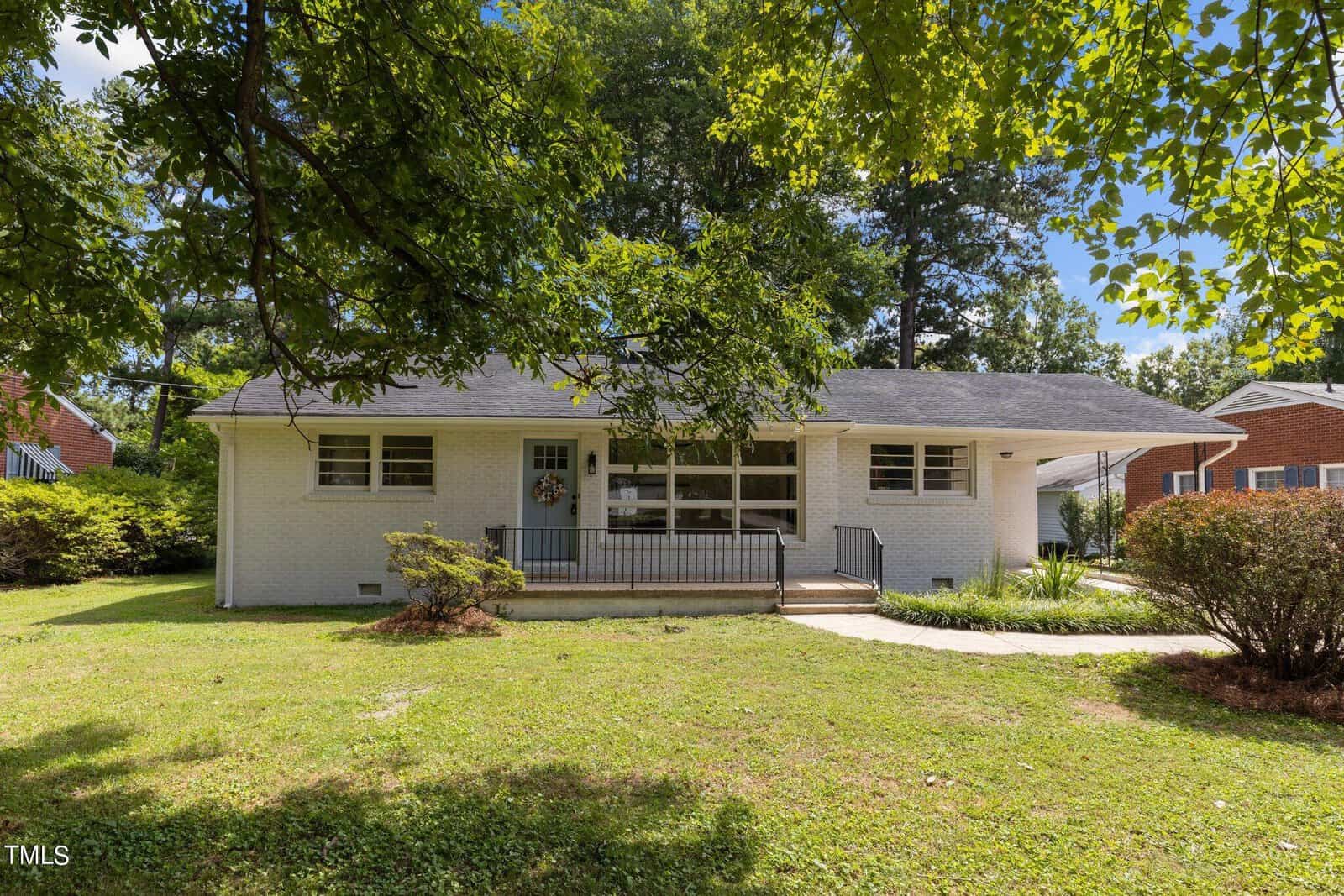 A single-story white brick house with a dark roof, surrounded by trees and greenery. The house has a small front porch with a wreath on the door, and a curved walkway leading to the entrance. The yard is grassy and bordered by shrubs.