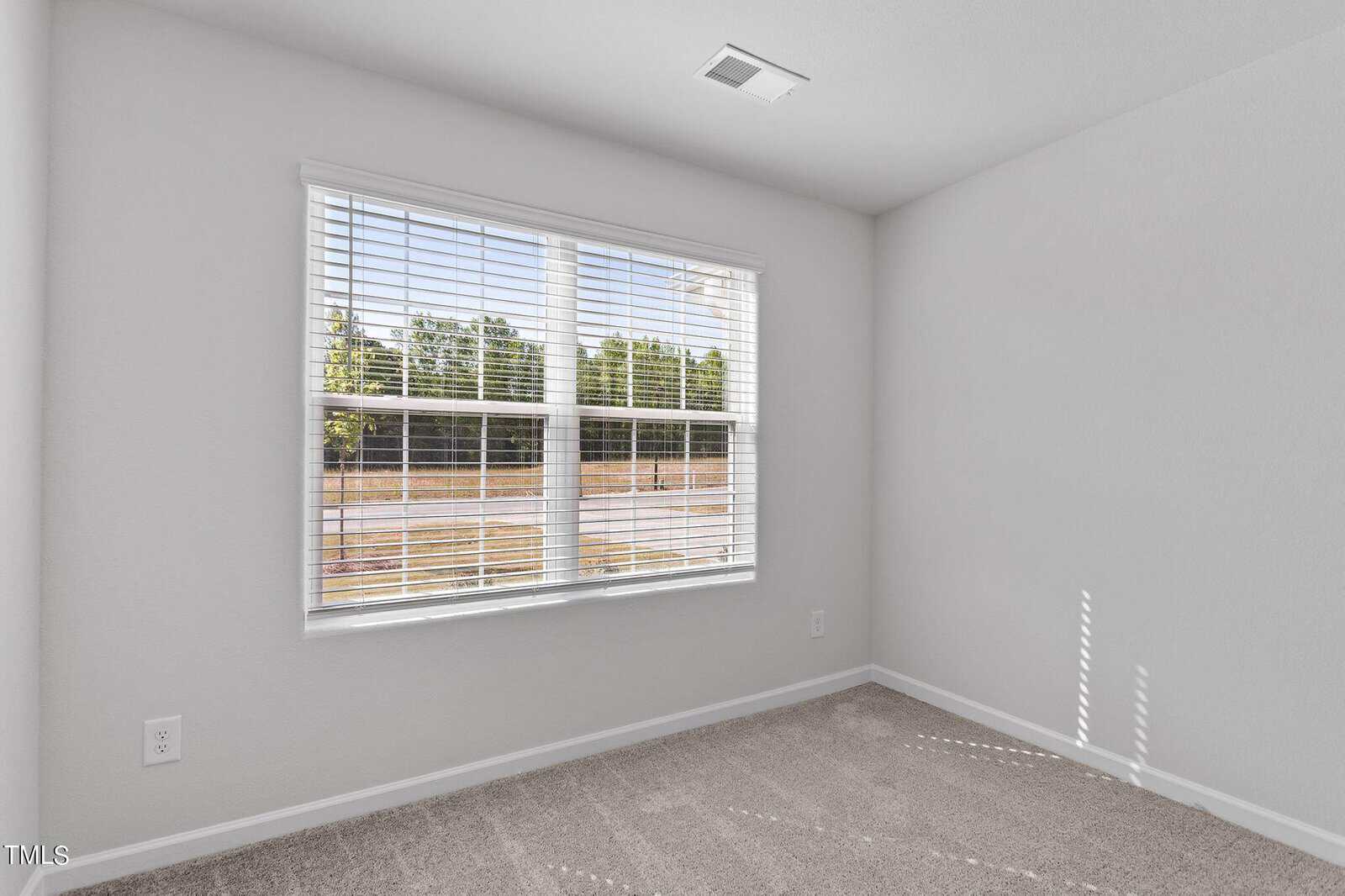 An empty room with light gray walls and a beige carpet. There is a large window with white blinds letting in natural light. Outside, trees and a fence can be seen. The room has a ceiling vent and a power outlet on the left wall.