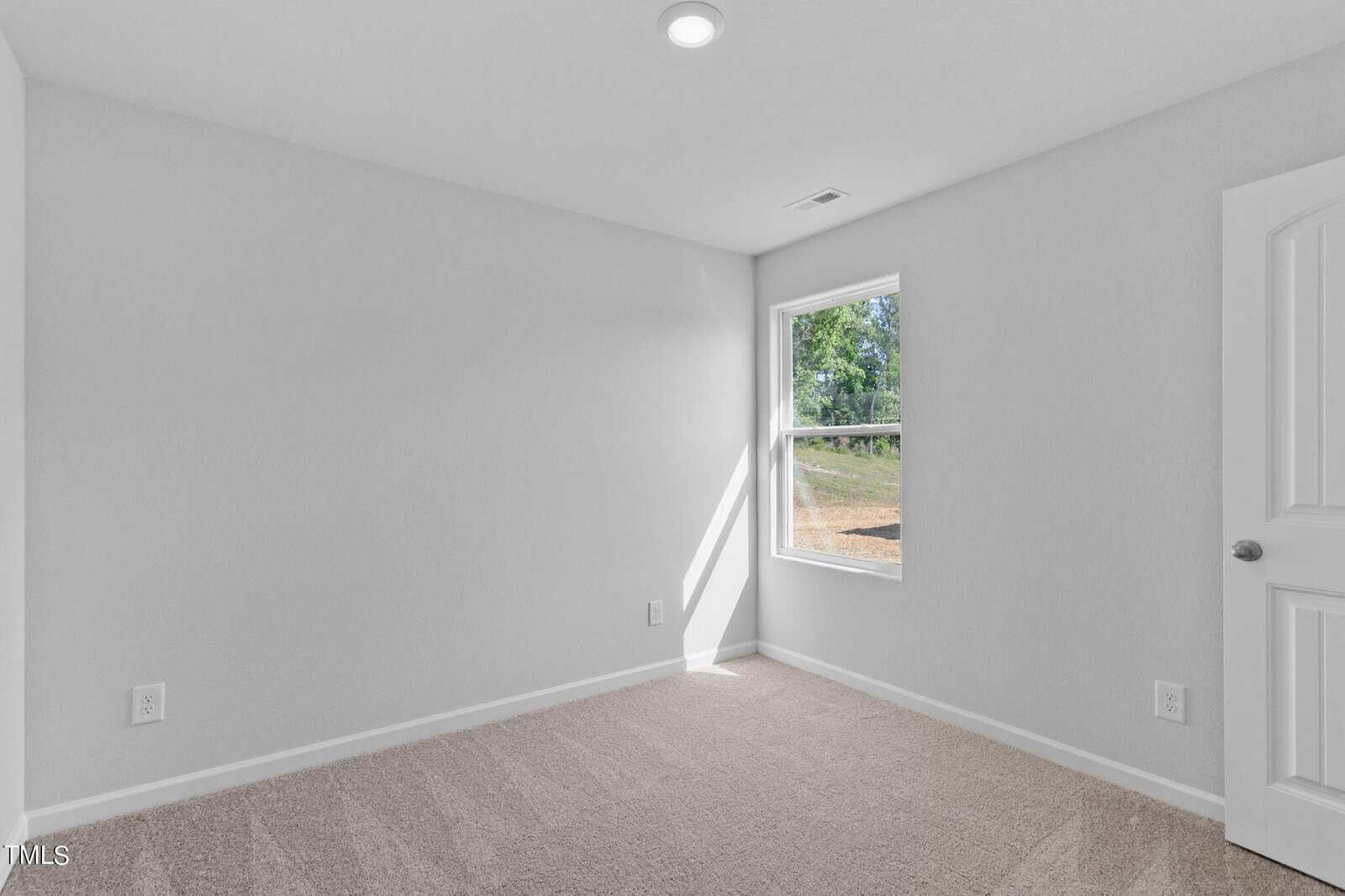 An empty room with light gray walls and a beige carpet. The room features a single window on one wall, allowing natural light to enter, and a closed white door on the right. A recessed ceiling light is turned on.