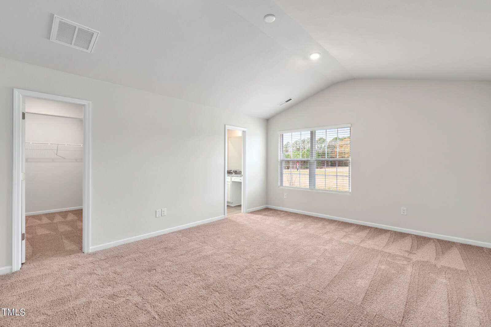 An empty room with beige carpet and light-colored walls, featuring a large window on the right side. There are doorways leading to a closet on the left and a bathroom in the center. The ceiling is vaulted with recessed lighting.