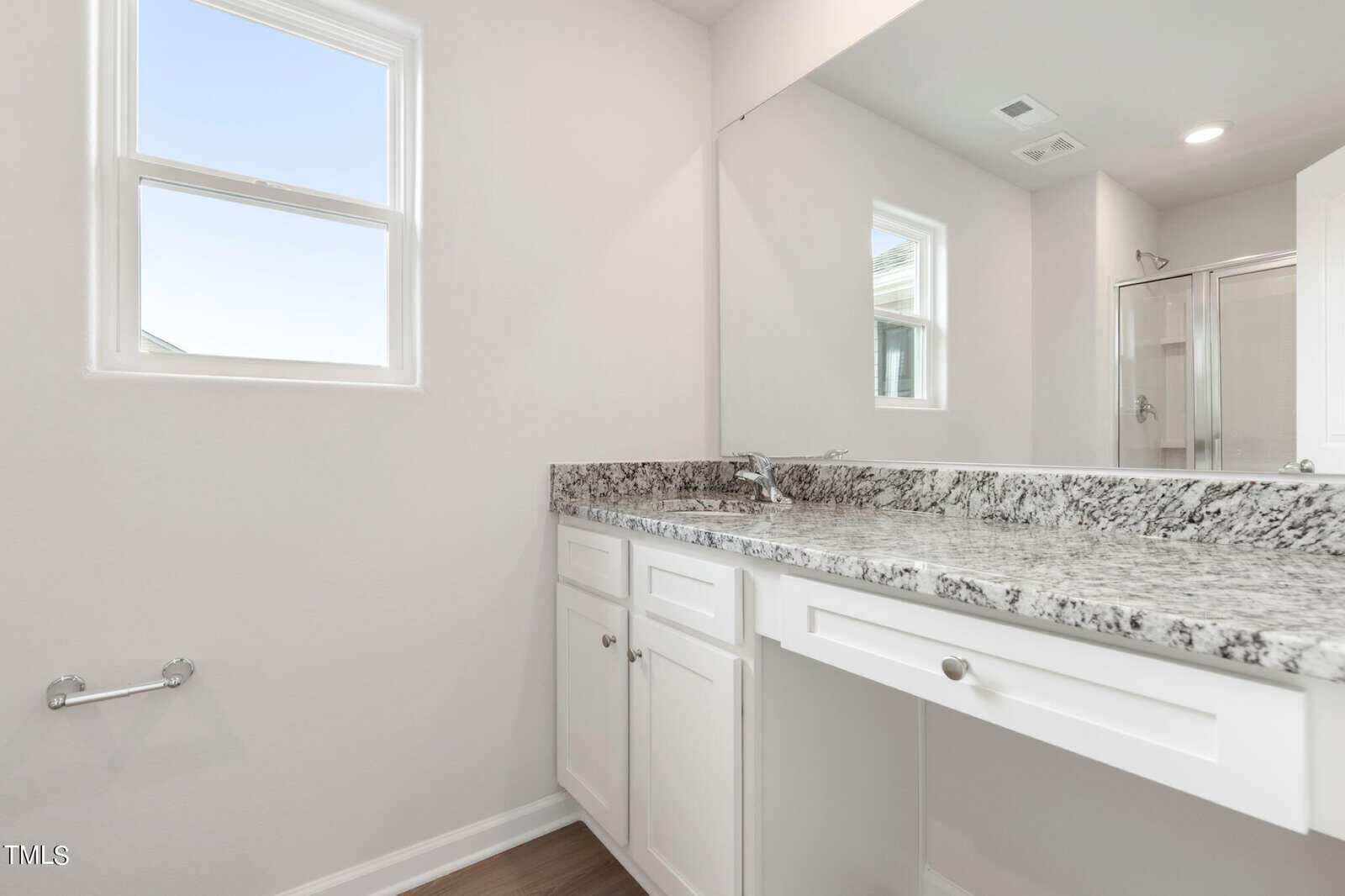 A bathroom with a speckled granite countertop, white cabinets, and a wall-mounted mirror. There are two small windows, a towel rack, and a shower with a glass door in the background. The walls are painted white.