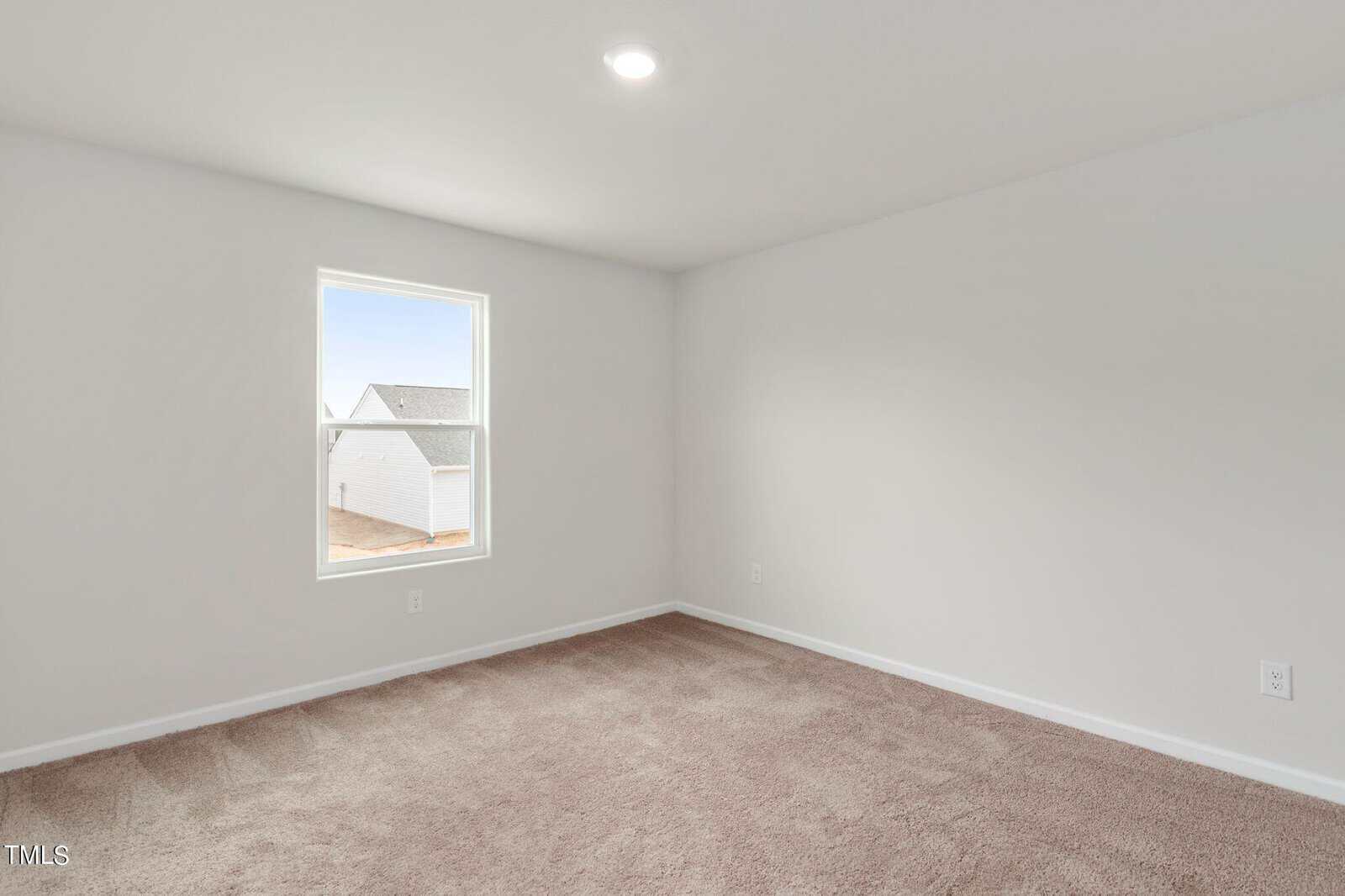 A bright, empty room with beige carpet, white walls, and a single window. A round ceiling light is switched on. The window offers a view of part of a neighboring building's roof and sky.
