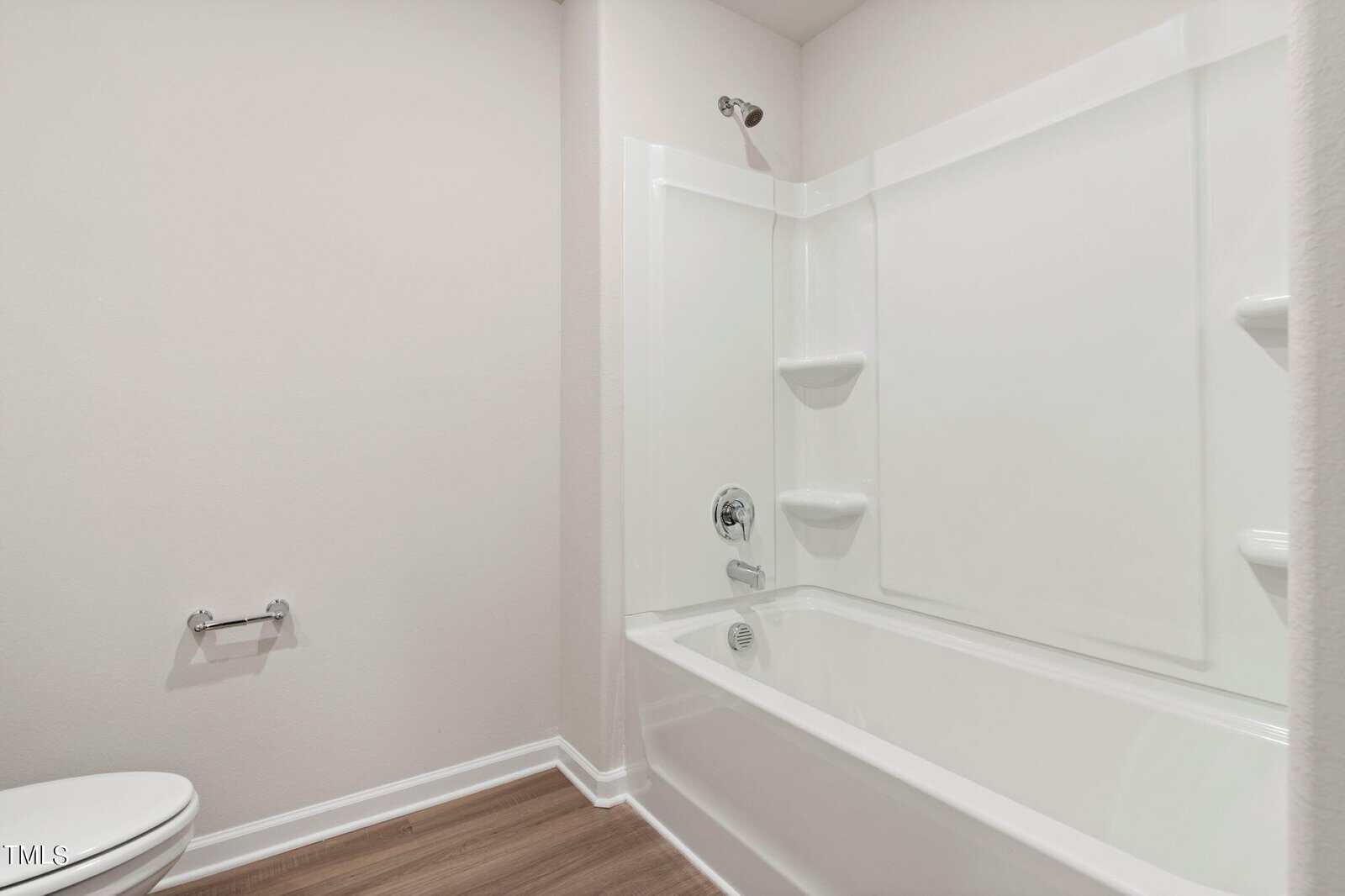 A bathroom featuring a white bathtub with a built-in shower and two corner shelves. The floor is covered with wood-patterned vinyl. A toilet paper holder is mounted on the left wall. The walls are painted in a light neutral color.