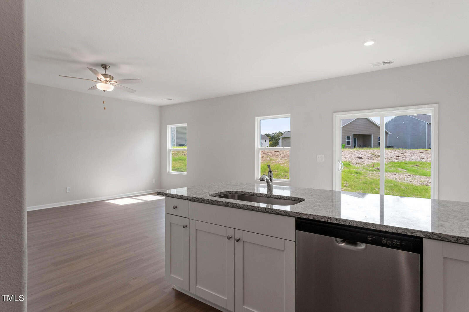 Open-concept kitchen and living area with light wood flooring. Features include a granite countertop with a sink and stainless steel dishwasher. Multiple windows provide natural light and offer views of a grassy landscape. Ceiling fan is visible.