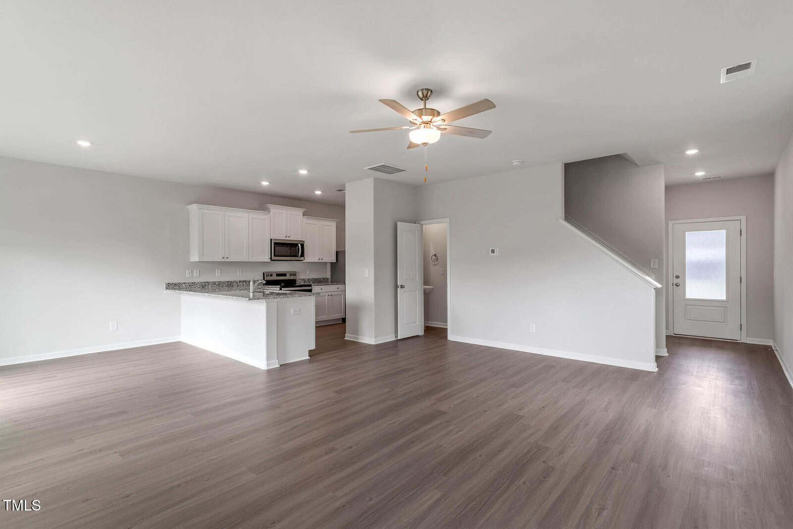 Empty living space with light wood flooring, white walls, and a ceiling fan. The room includes a modern kitchen with white cabinets, granite countertops, and stainless steel appliances. A door and staircase are visible in the background.