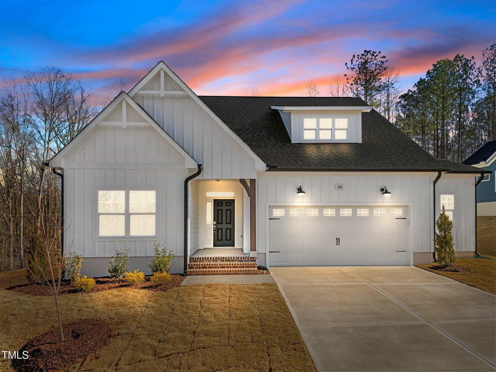 A modern single-family house with white siding, black roof, double garage, and illuminated windows sits on a freshly landscaped yard at sunset with a colorful sky in the background.