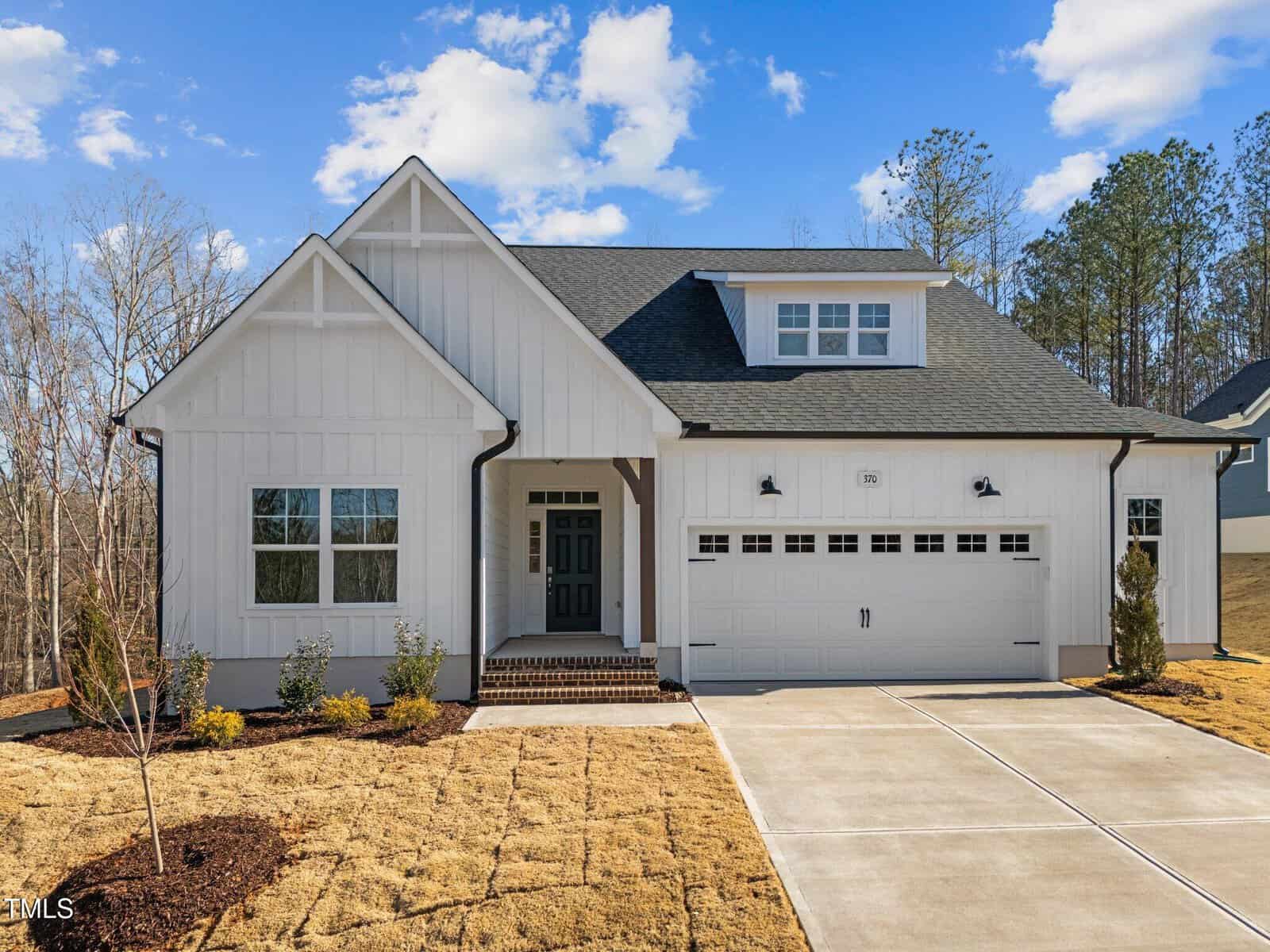 A modern white house with board and batten siding, a peaked roof, double garage, front porch, and landscaped yard with small shrubs, set against a backdrop of trees and a blue sky.