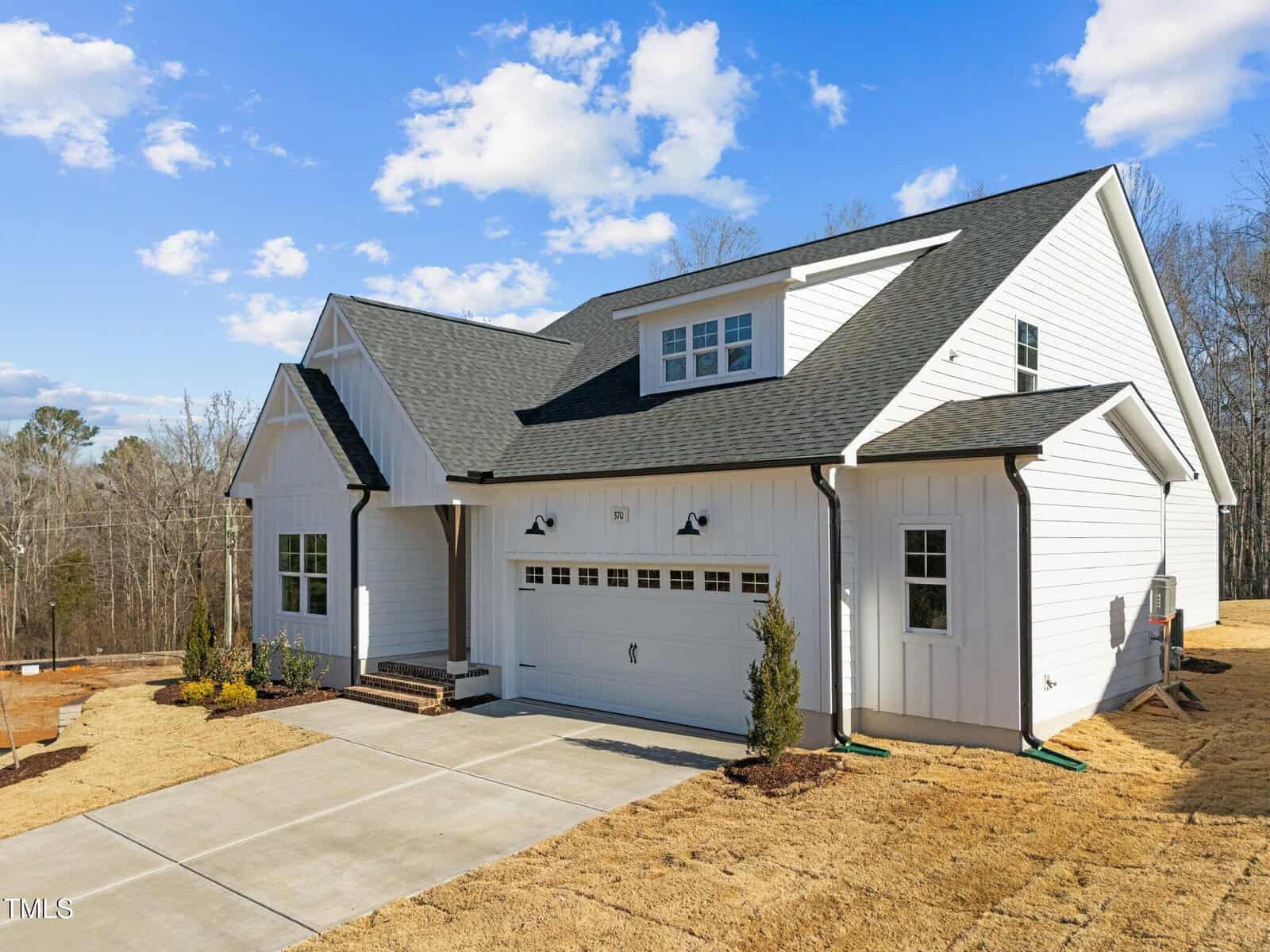 A modern, white two-story house with black roof shingles, a double garage, and a driveway. The yard is covered with straw and a few young shrubs. The sky is partly cloudy and trees are in the background.