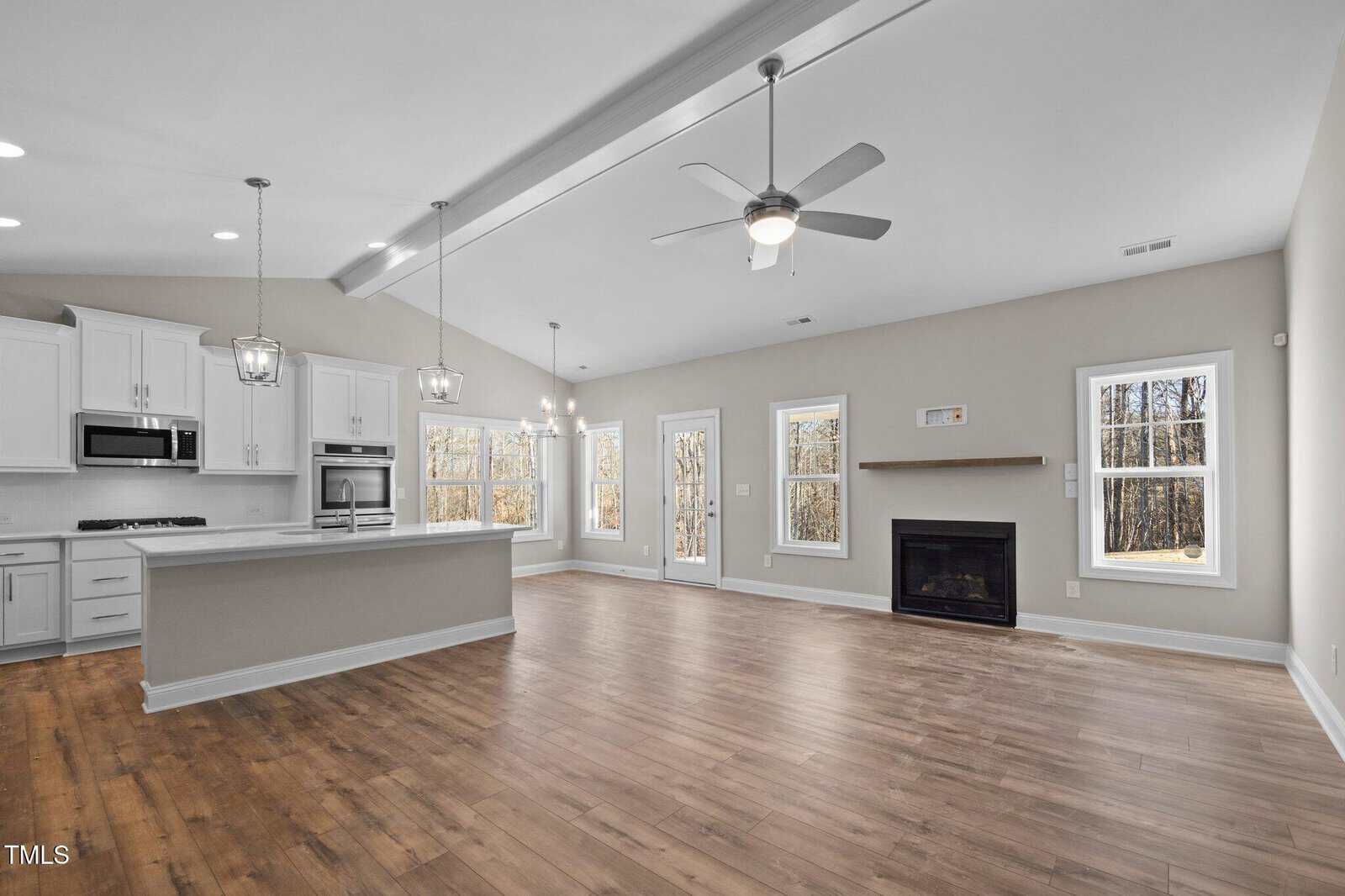 Open-concept living area with wood floors, white kitchen featuring an island, stainless steel appliances, and pendant lights, plus a living space with a ceiling fan, fireplace, and large windows.