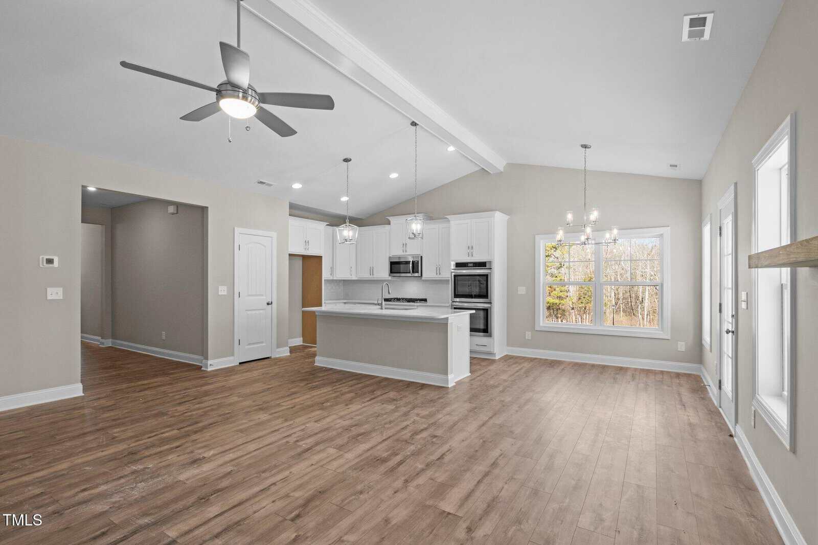 Open-concept kitchen and living area with light wood floors, white cabinets, stainless steel appliances, center island, and large windows letting in natural light; ceiling fans and pendant lights are visible.