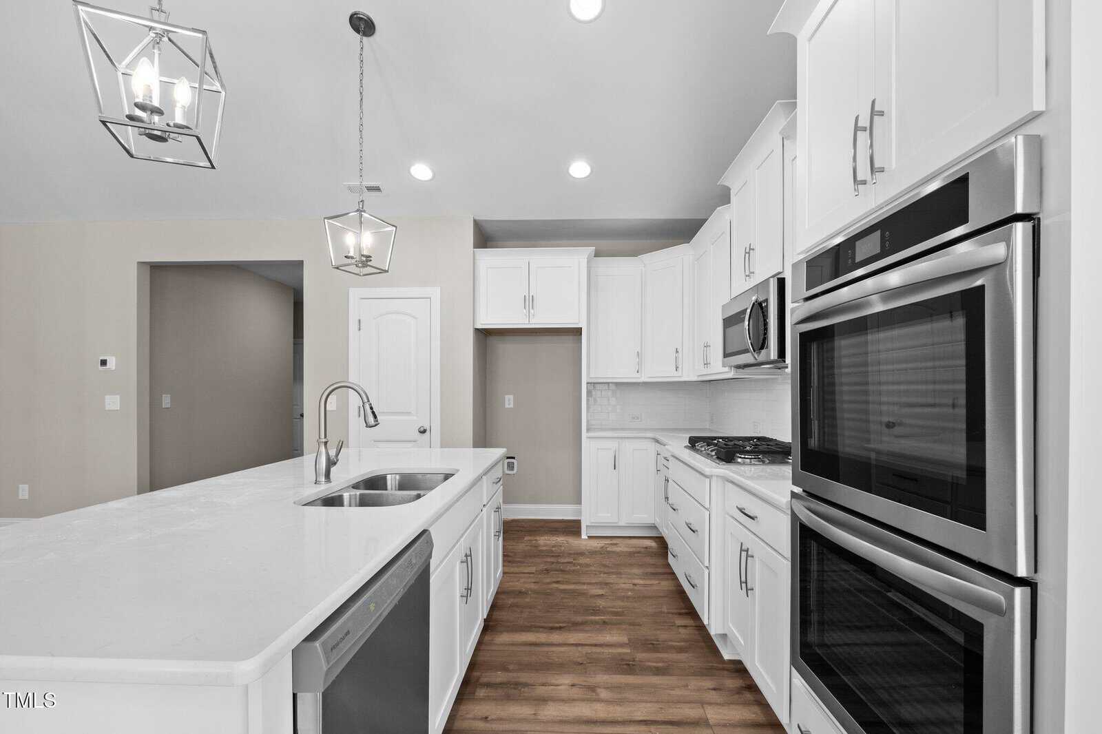 A modern kitchen with white cabinets, stainless steel appliances, a built-in oven, a microwave, a double sink in a large island, pendant lights, and wood flooring. The walls are light and the space appears well-lit.