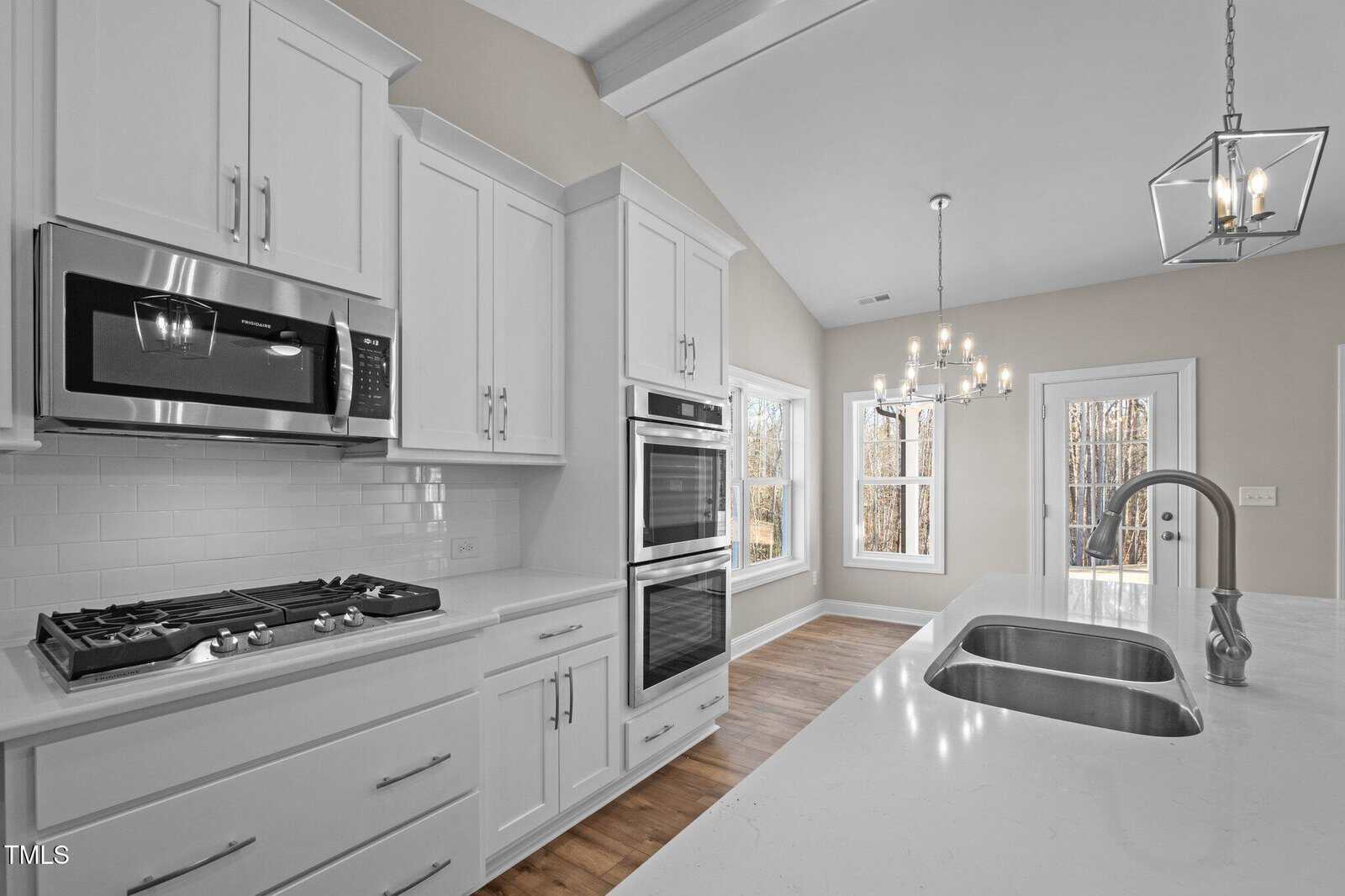 A modern kitchen with white cabinets, stainless steel appliances, a gas cooktop, double wall ovens, a microwave, a large island with a sink, and pendant lighting. A dining area with large windows is visible in the background.