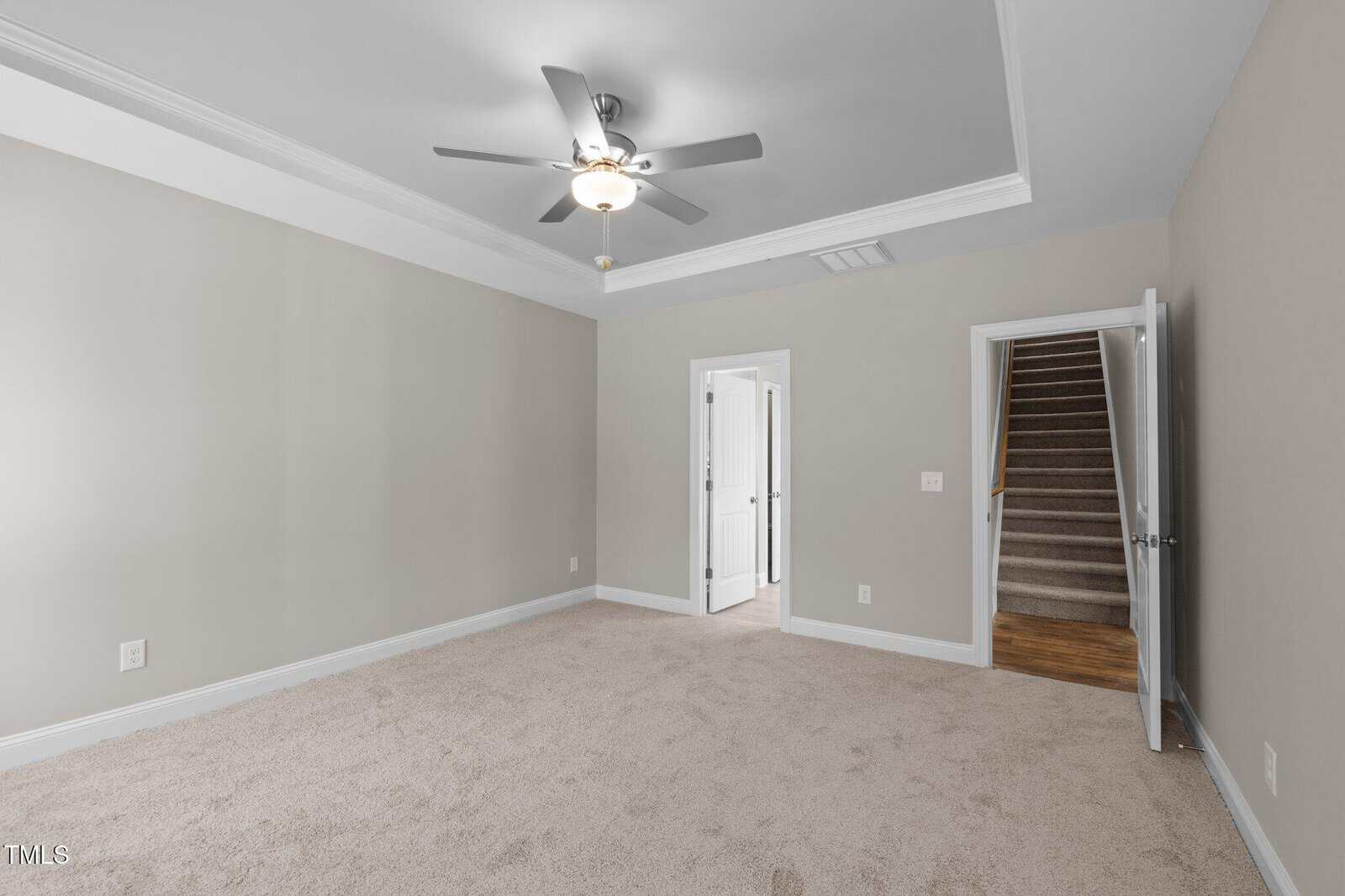 Empty carpeted room with light gray walls, white trim, and a ceiling fan with lights. There is a door leading to another room, an open door to a stairway, and a white vent on the wall near the ceiling.