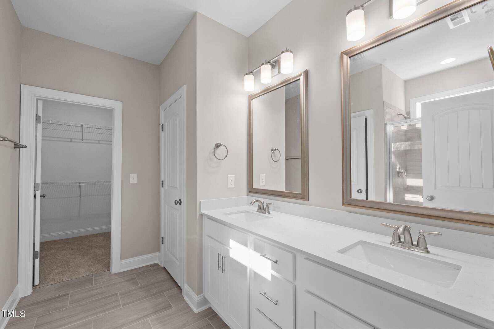 Modern bathroom with double sinks, white cabinets, two mirrors, and wall-mounted lights. A walk-in closet with wire shelves is visible through an open door on the left. Neutral colors and clean lines throughout.
