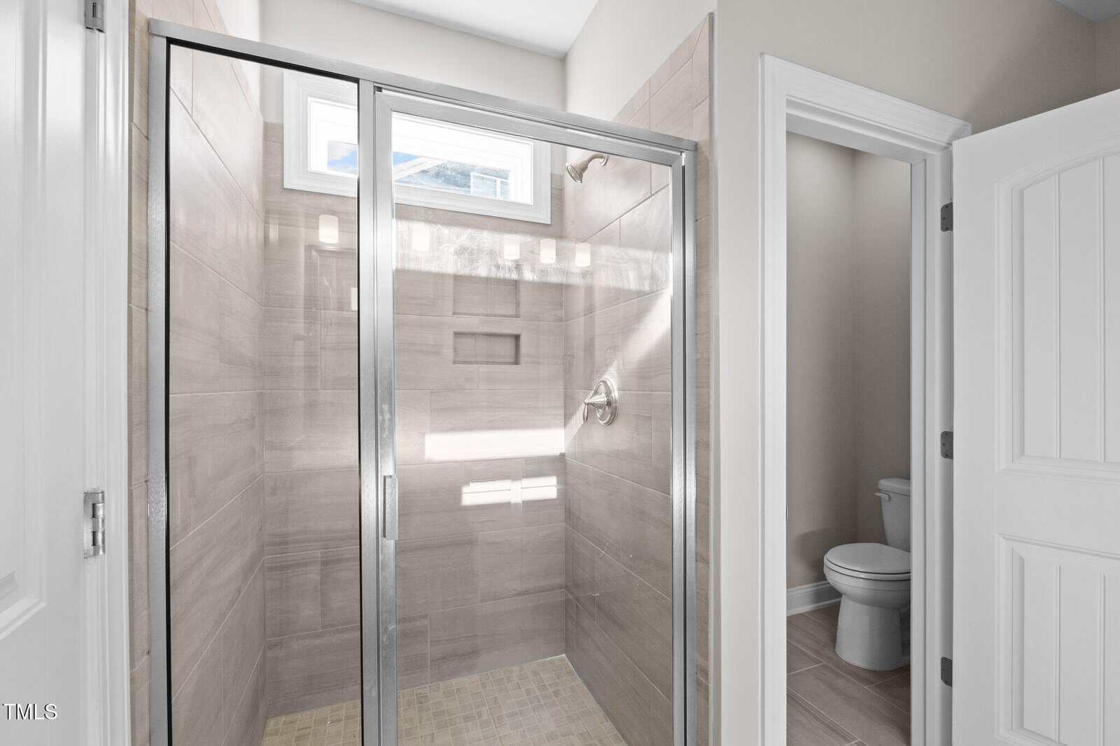 A bathroom with a glass-door shower featuring beige tiles and a built-in shelf. A small window above lets in light. A separate toilet room is visible through an open door.