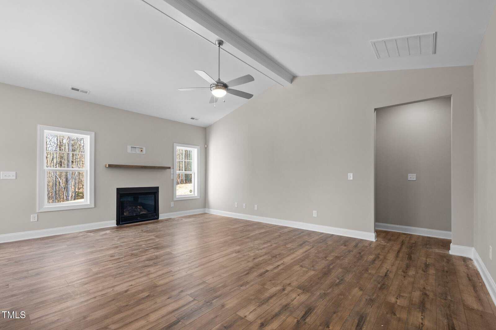 A spacious, empty living room with light beige walls, wood flooring, a ceiling fan with light fixture, two windows, a fireplace with a wood mantel, and an arched doorway leading to another room.