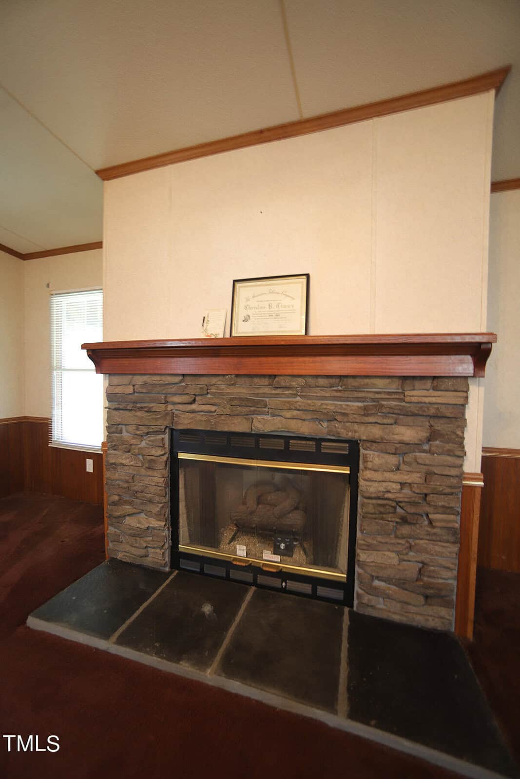A brick fireplace with a dark stone hearth and a wooden mantel. On the mantel are a framed certificate and a small decorative item. The room has light-colored walls and dark wood paneling below the windowsill. The carpet is a dark, rich brown.
