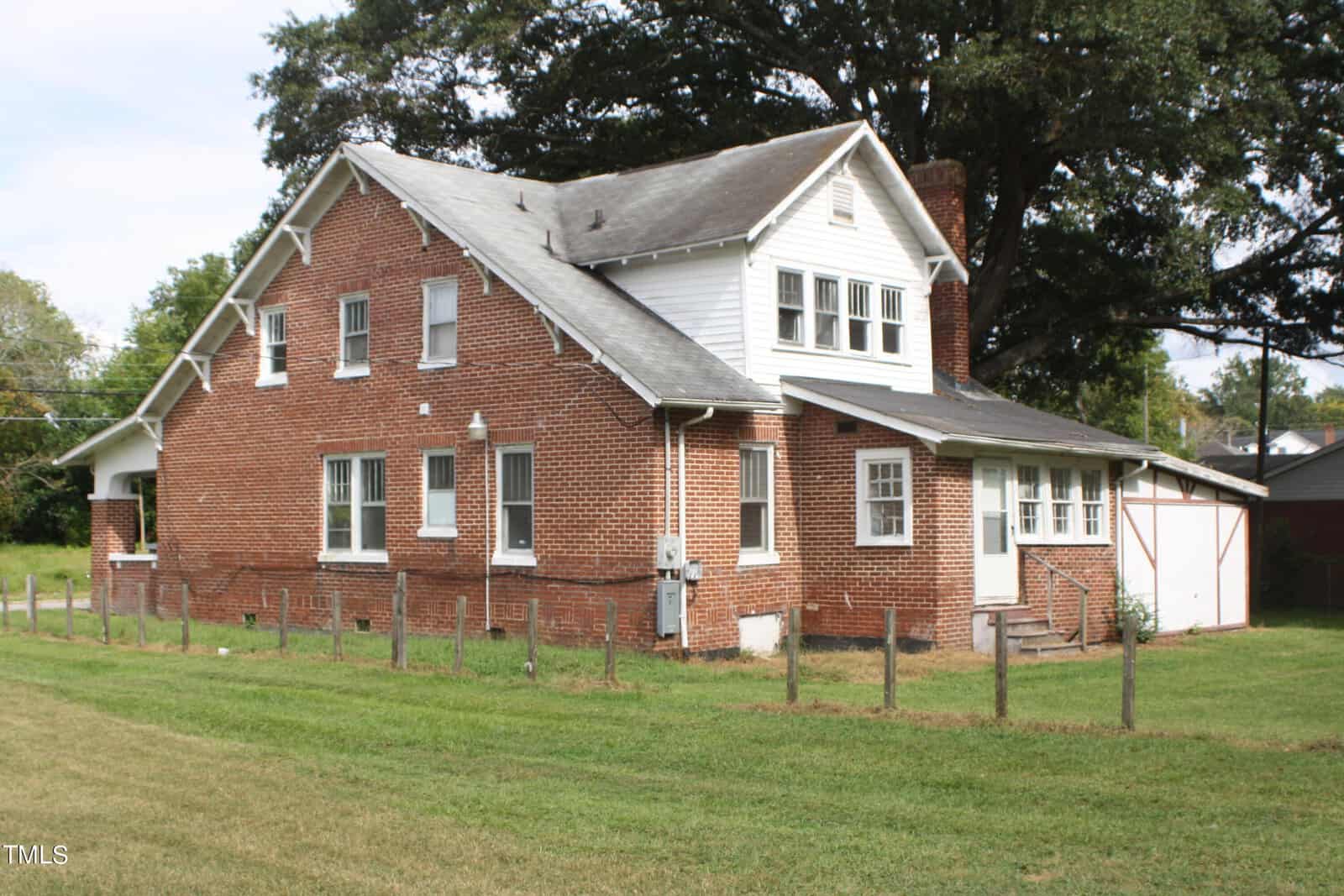 A two-story red brick house with a gabled roof and white trim. The house features multiple windows, a chimney, and a small porch area at the back. A large tree stands behind the house, and the yard is mostly grass with a small fence on one side.