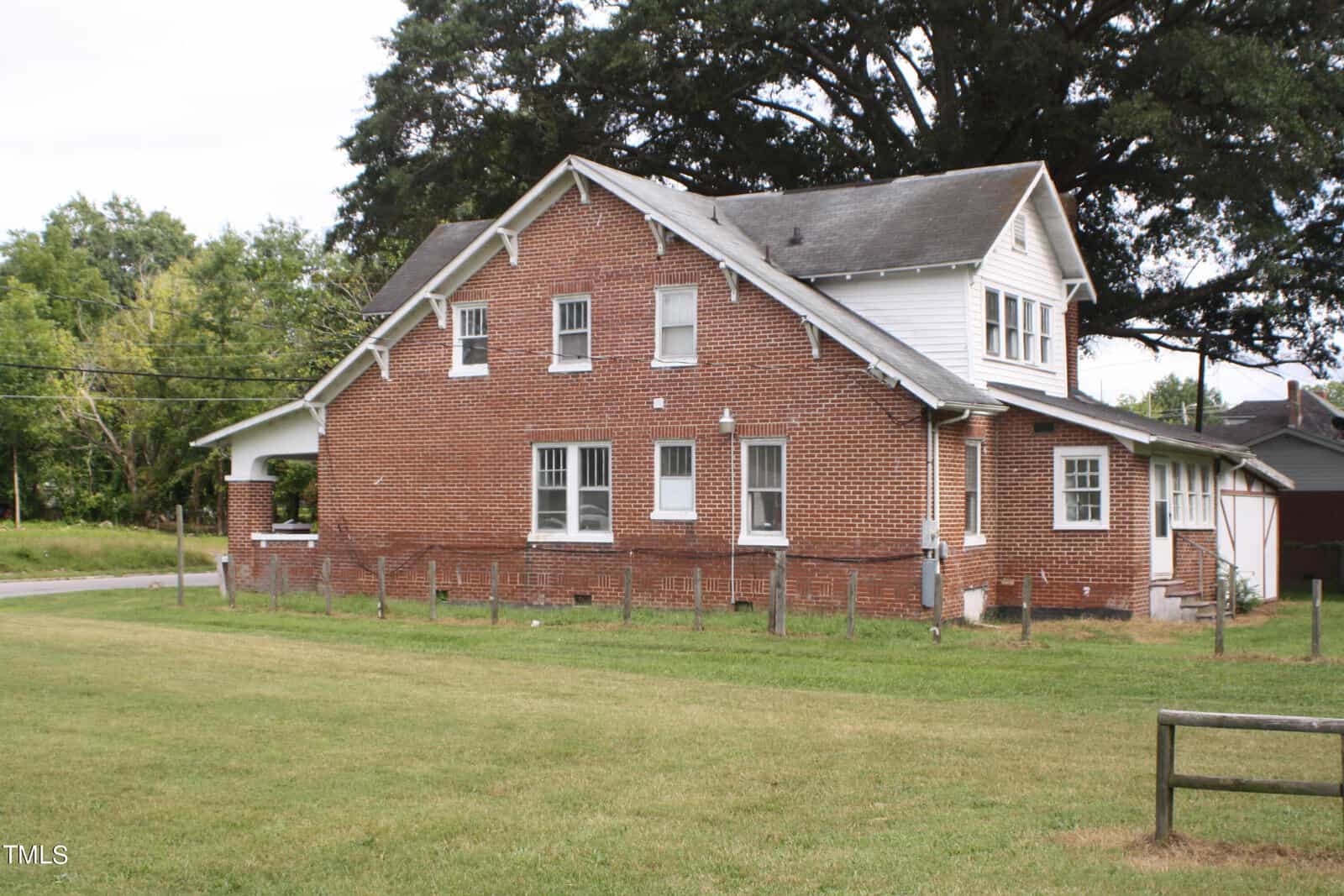 A two-story brick house with white-trimmed windows and a gabled roof stands in a grassy yard. An adjacent wooden fence partially surrounds the property. Trees and a paved road are visible in the background.