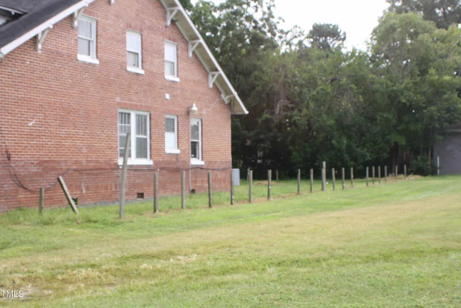 A red brick building with several windows stands on the left side of the image. A grassy area extends in front of the building, bordered by a low wooden fence. Trees and greenery are visible in the background. The sky is overcast.