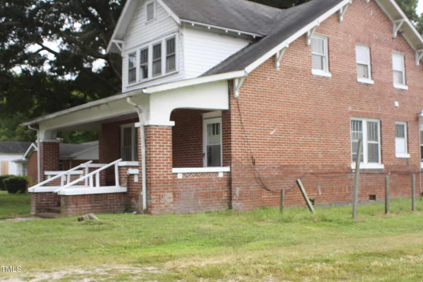 A two-story brick house with a gabled roof, white trim, and a covered front porch with a white railing. The house has several windows and an adjacent grassy yard. A few trees are in the background. The building appears older, showing some wear.