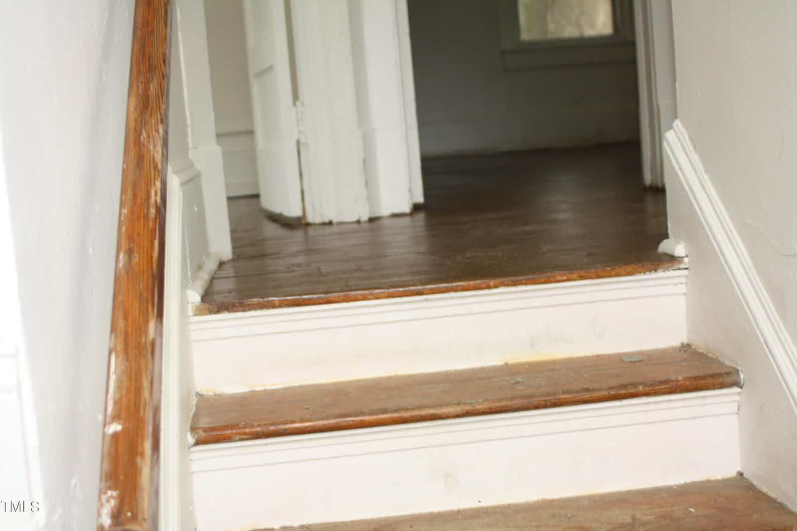 A close-up view of an indoor wooden staircase with three steps leading to a hallway. The steps have visible wear, and the white walls show slight discoloration. A wooden handrail is mounted to the left side. An open door and a window are visible in the background.