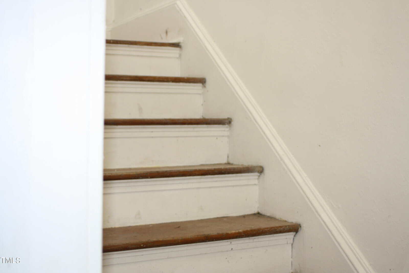 An interior shot of a narrow wooden staircase with white-painted risers and brown treads. The steps show signs of wear, and marks are visible on the paint. A white wall with subtle texture lines the right side of the staircase.