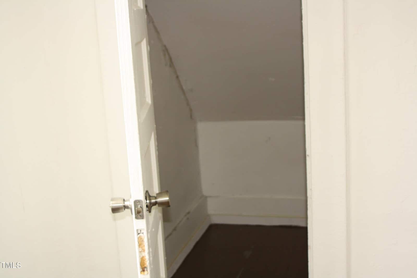 A small, empty room with white walls, a dark brown floor, and a slanted ceiling is seen through a partially open white door with a silver doorknob. The door frame shows signs of wear.
