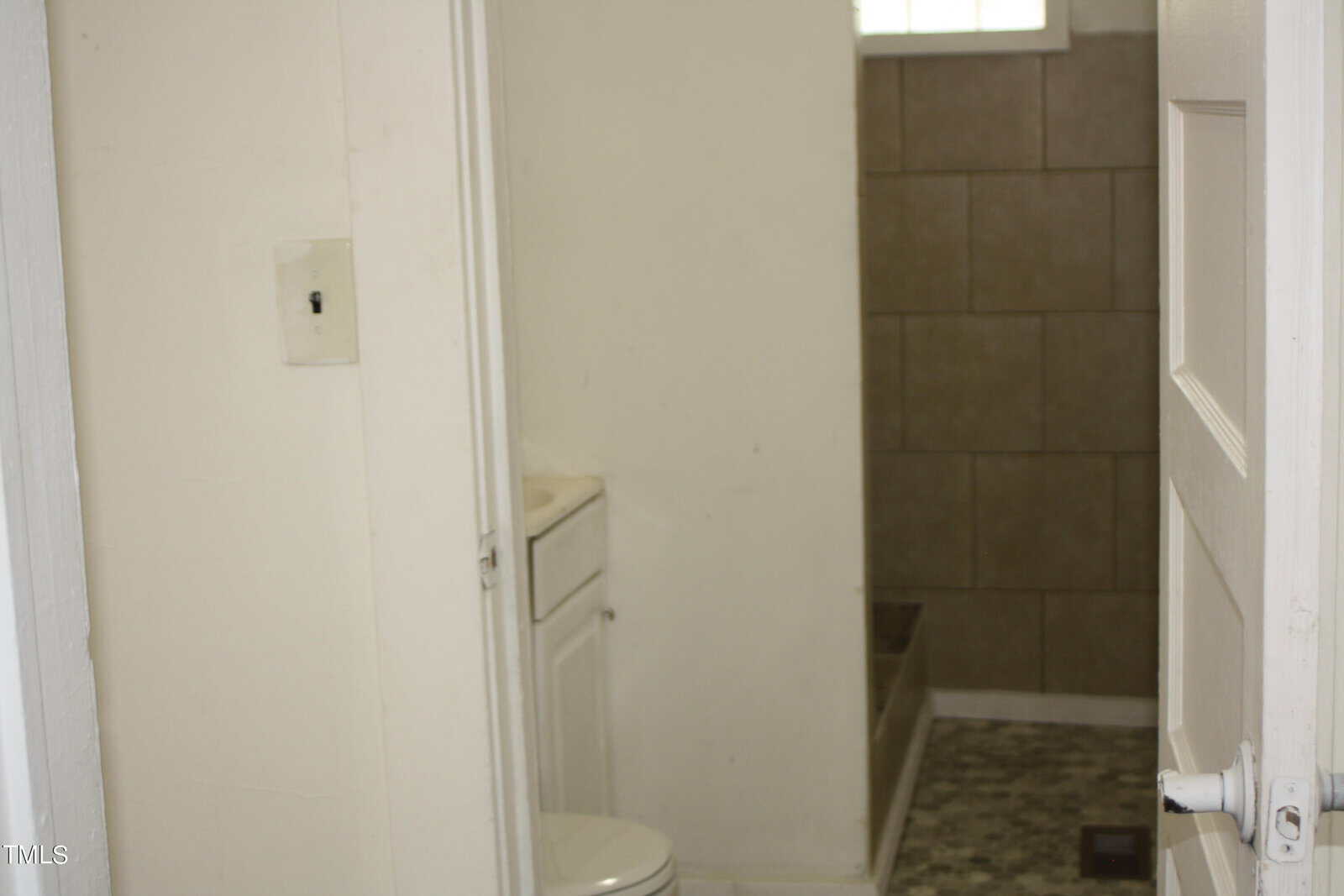 A small bathroom with a partially open door showing a white toilet and a white vanity against the wall. There is a tiled shower area with brown tiles and a small window above. The walls are white and the floor has a mosaic tile pattern.