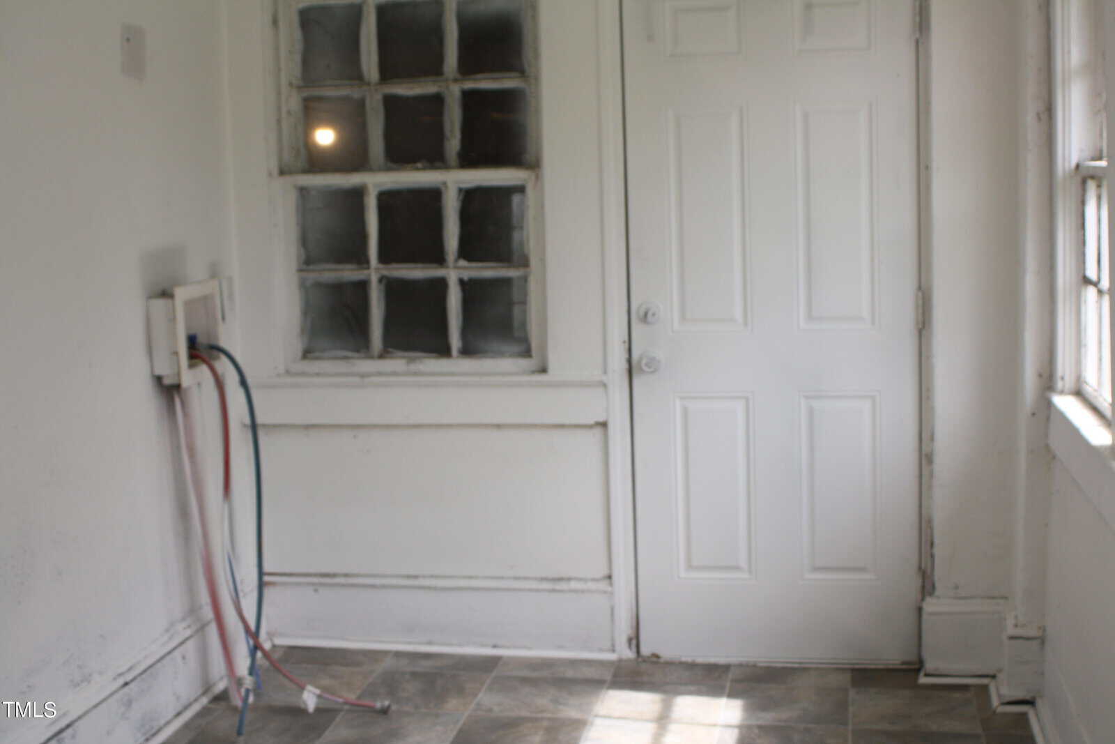 A small, empty room with white walls and a white door. There is a set of tangled wires and hoses attached to the wall on the left. The floor is covered with grey tiles, and a single window with a grid pattern is on the left side of the door.