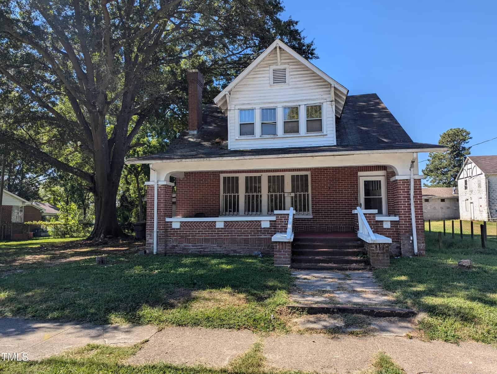 A two-story brick house with a covered porch and white trim stands on a sunny day at 219 W Rockspring Street, Henderson. Large windows grace both upper and lower floors. A tall tree to the left casts shade over part of the house and lawn, while a concrete walkway leads to the porch stairs.