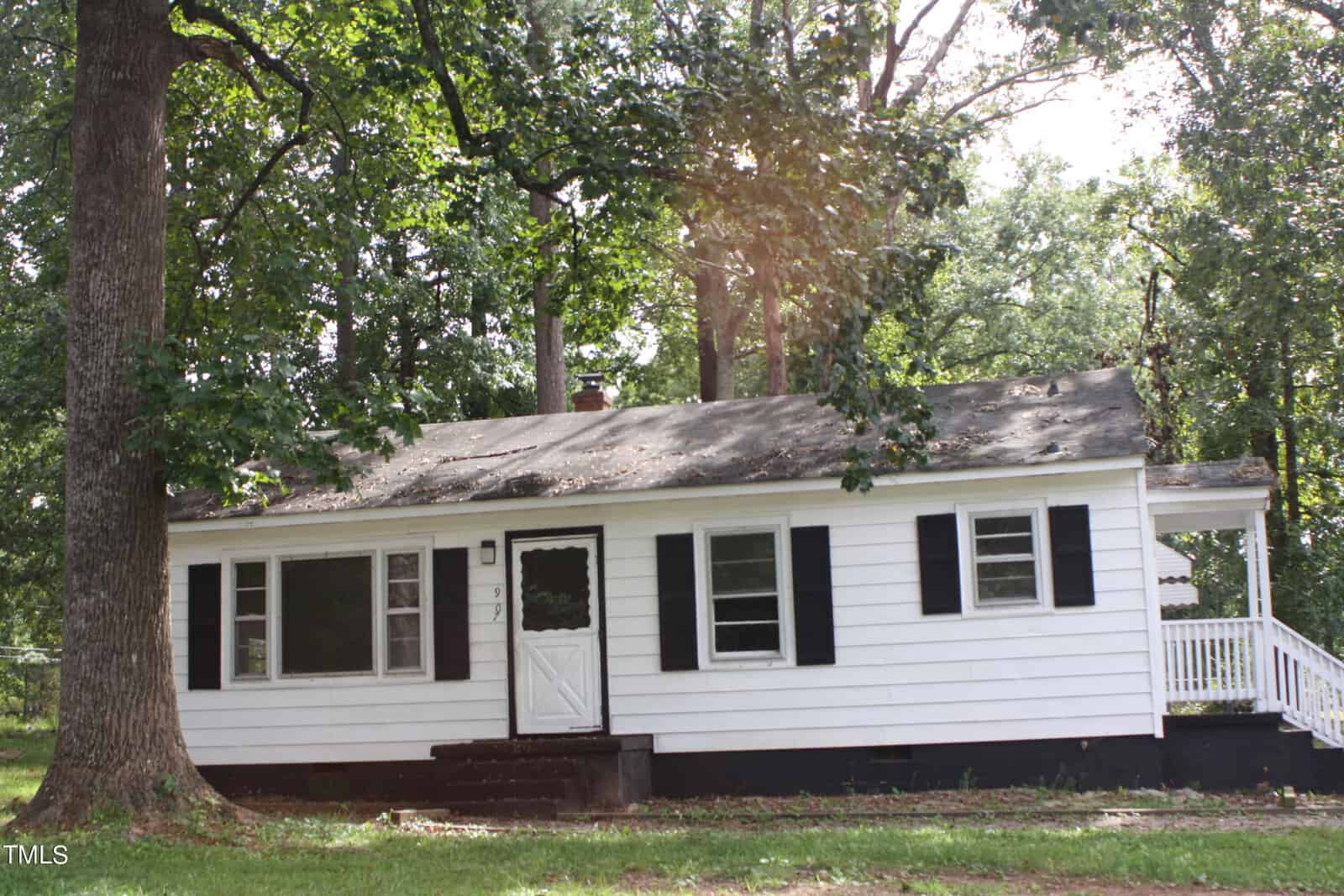 A small white house with black shutters at 907 Abbott Street in Henderson, partially shaded by large trees. The home features a front door with a black decorative glass panel, two front windows, and an accessible side ramp leading to the entrance. The lawn around the house is green and lush.