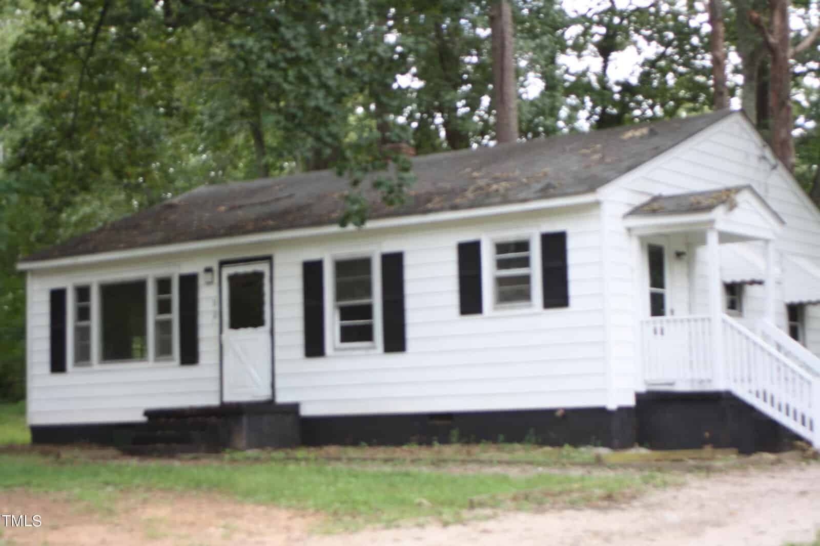 A small white house with black shutters and a front porch is surrounded by trees. The house has a dark foundation and white steps leading up to the front door. The surrounding grassy area shows a dirt path leading to the house.