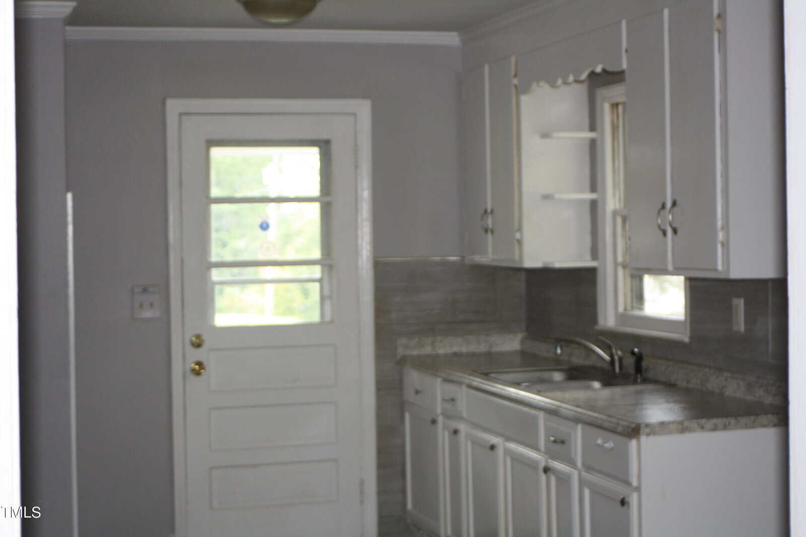 A small kitchen with gray walls and cabinets. It features a sink under a window, a countertop, and a door with a window panel leading outside. The kitchen has a gray tiled backsplash and white trim.