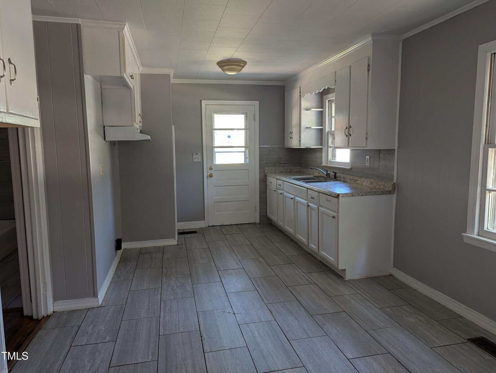 A clean, empty kitchen with light gray walls and large gray floor tiles. It features white cabinets, a sink under a window, and a back door with a glass panel. The area is well-lit with natural light from the windows. There's a ceiling light fixture in the center.