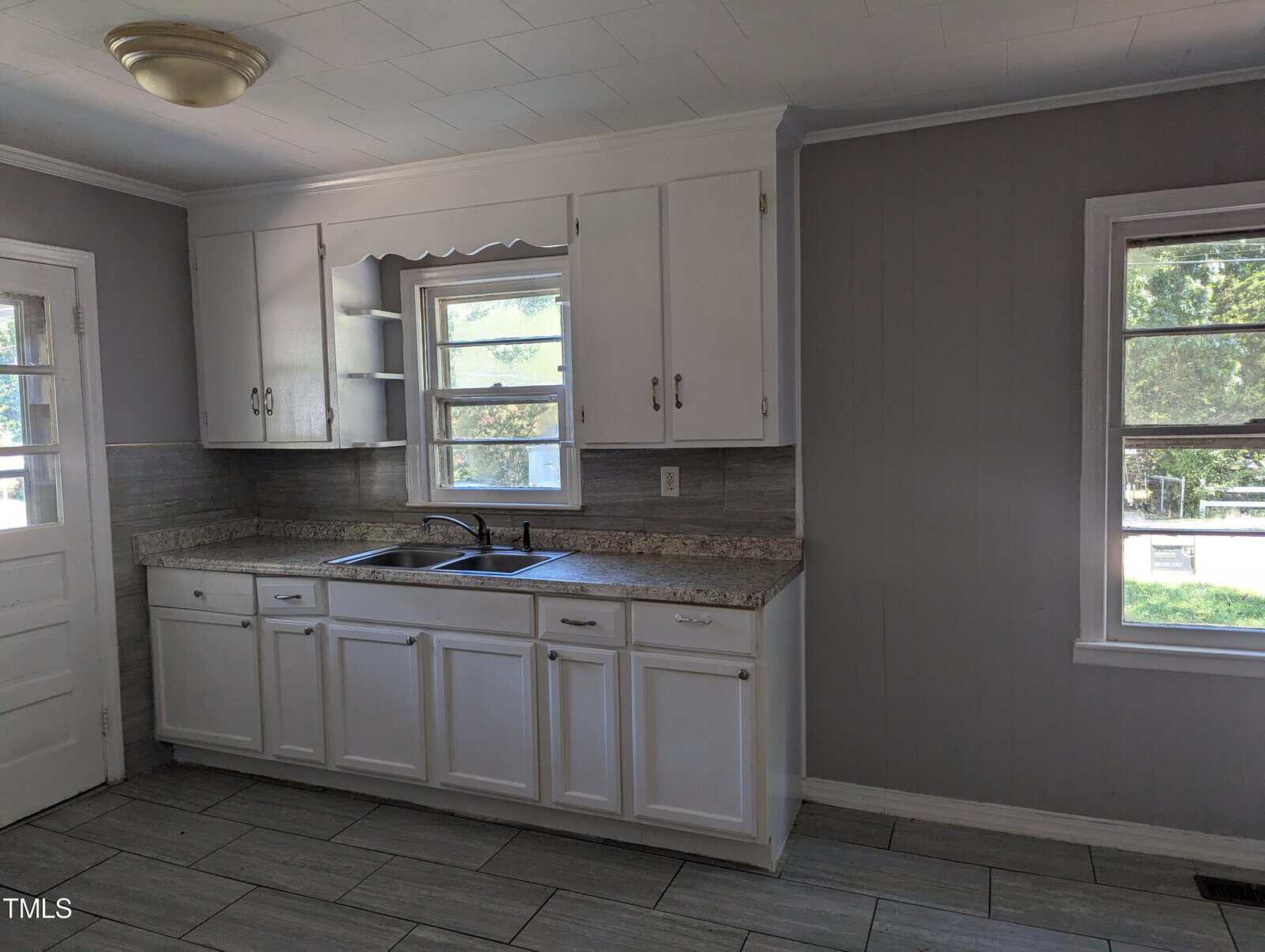 A kitchen with white cabinetry, a double sink under a window, granite countertops, and gray tile flooring. Natural light streams in through two windows, highlighting the clean, minimalist space. A door is visible on the left side of the room.