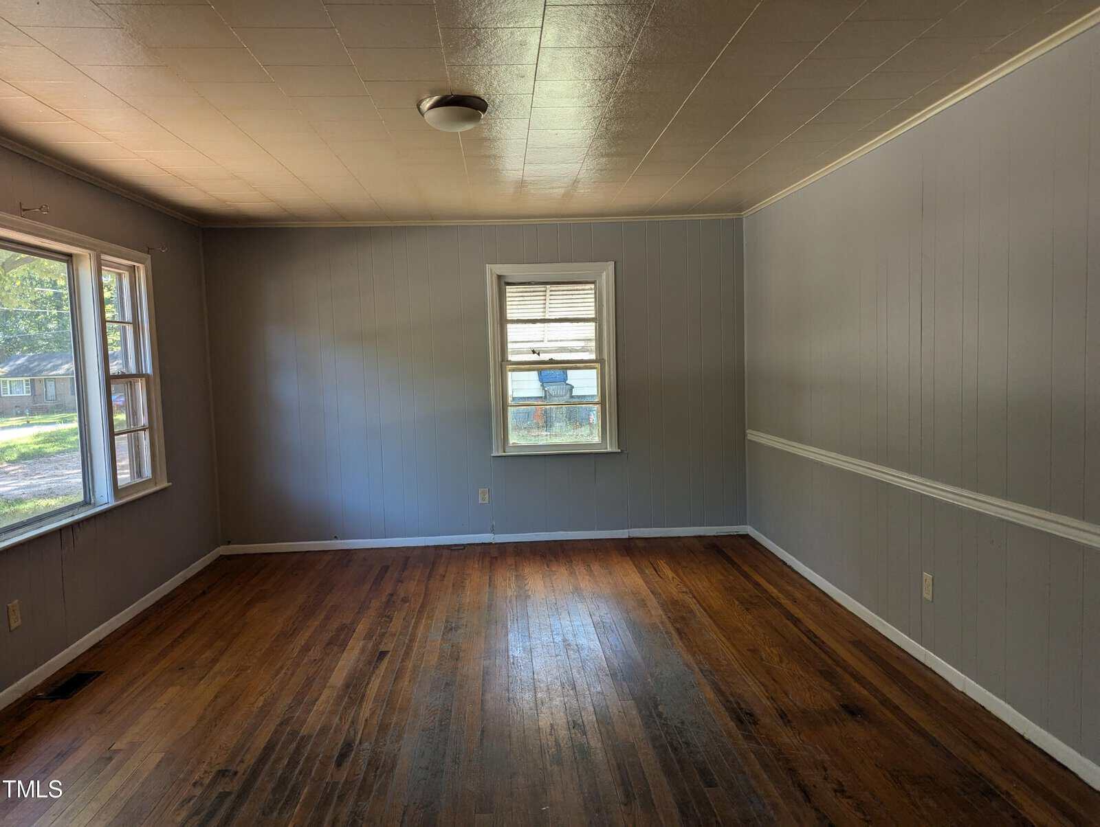 An empty room with light gray paneled walls, a wooden floor, and a white ceiling. The room has three windows, with one on the far wall and two on the adjacent wall, letting in natural light. A ceiling light fixture is visible, and there's white molding along the walls.