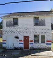 The two-story white brick building at 610 N Chestnut Street, with its red door and accents of red and blue horizontal stripes, features a single-car garage on the left side and two windows on the right. The exterior appears weathered and worn, with dark stains near the roof in Henderson.
