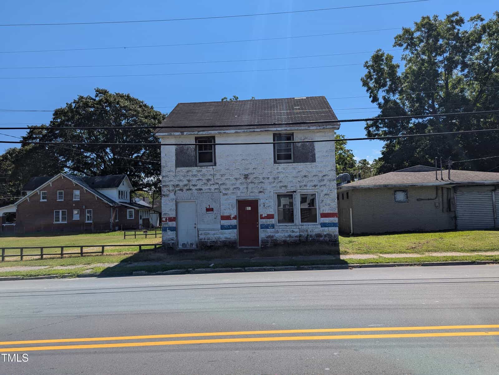 A small, weathered, two-story white building with a red door and a faded blue and red stripe along the front. It stands adjacent to a single-story green building and a red brick house in the background at 610 N Chestnut Street in Henderson. Power lines cross the clear blue sky above the road in the foreground.