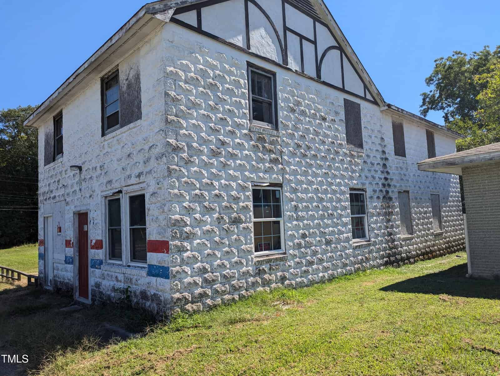 A two-story, stone-clad building with a white exterior, patches of red and blue paint, and multiple boarded-up windows. There's a small red door on the left and a grassy area surrounding the building. The sky is clear and blue.