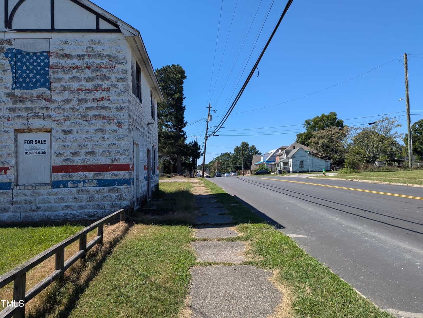 A rural street scene featuring an old stone building with a faded American flag painted on the side. A "For Sale" sign is visible on the building. There's a paved road with a few houses and trees lining the street on a sunny day.