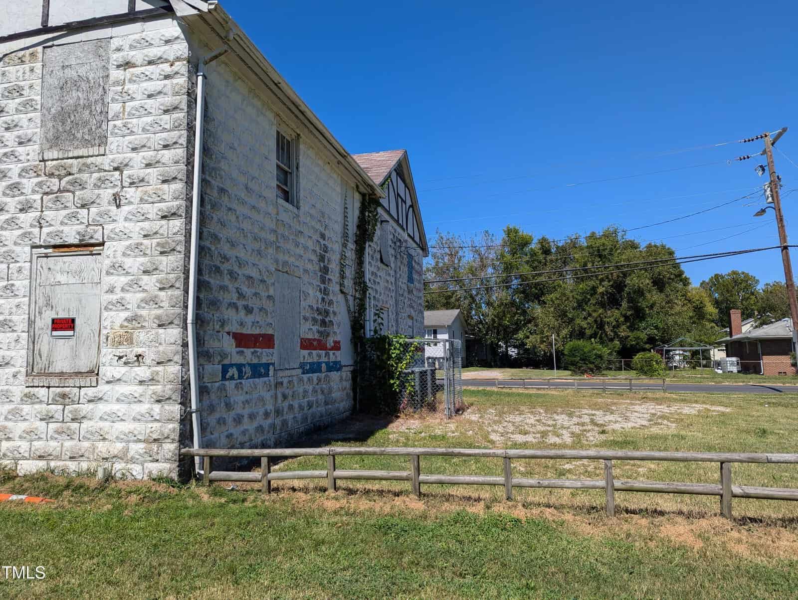 An old, abandoned stone building with boarded-up windows and creeping ivy on the side. It sits on a grassy lot with a small wooden fence in front. A chain-link fence is visible in the background, along with power lines, trees, and other buildings under a clear blue sky.