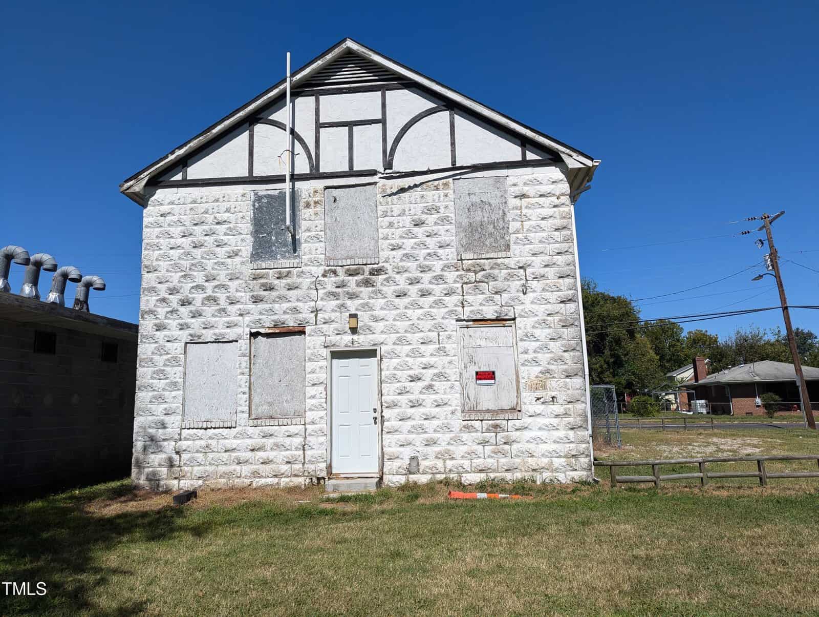 A two-story, white stone building with boarded-up windows and a single door in the center. The building has a neglected appearance and is set against a clear blue sky. In the background, there are power lines, a grassy yard, and neighboring houses.