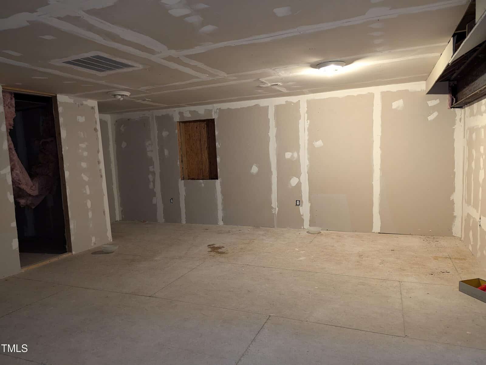 A partially finished basement with drywall panels in place but not yet painted. Exposed insulation is visible through an open doorway on the left. The floor is bare, and the lighting consists of a single ceiling light fixture. There is an unfinished window opening.