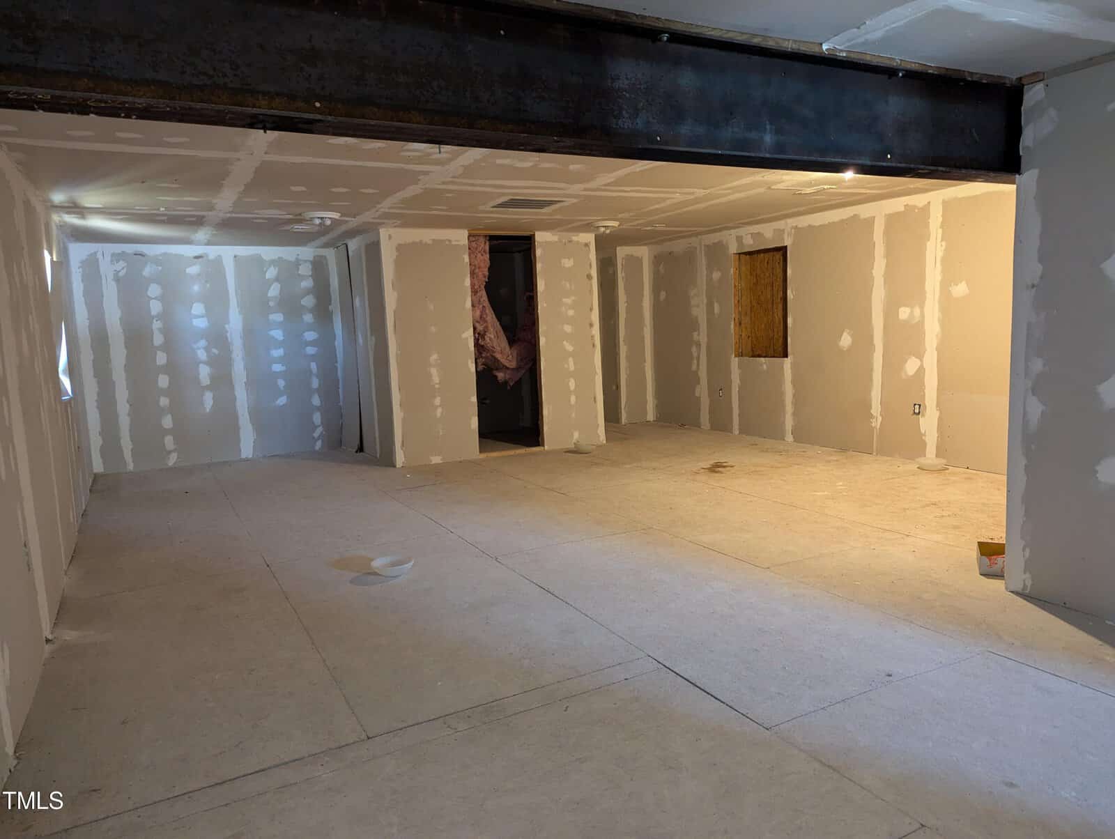 A room under construction with unfinished drywall and exposed insulation. The floor is covered with construction materials. There is a large steel beam running across the ceiling and a partially installed window on the right wall.